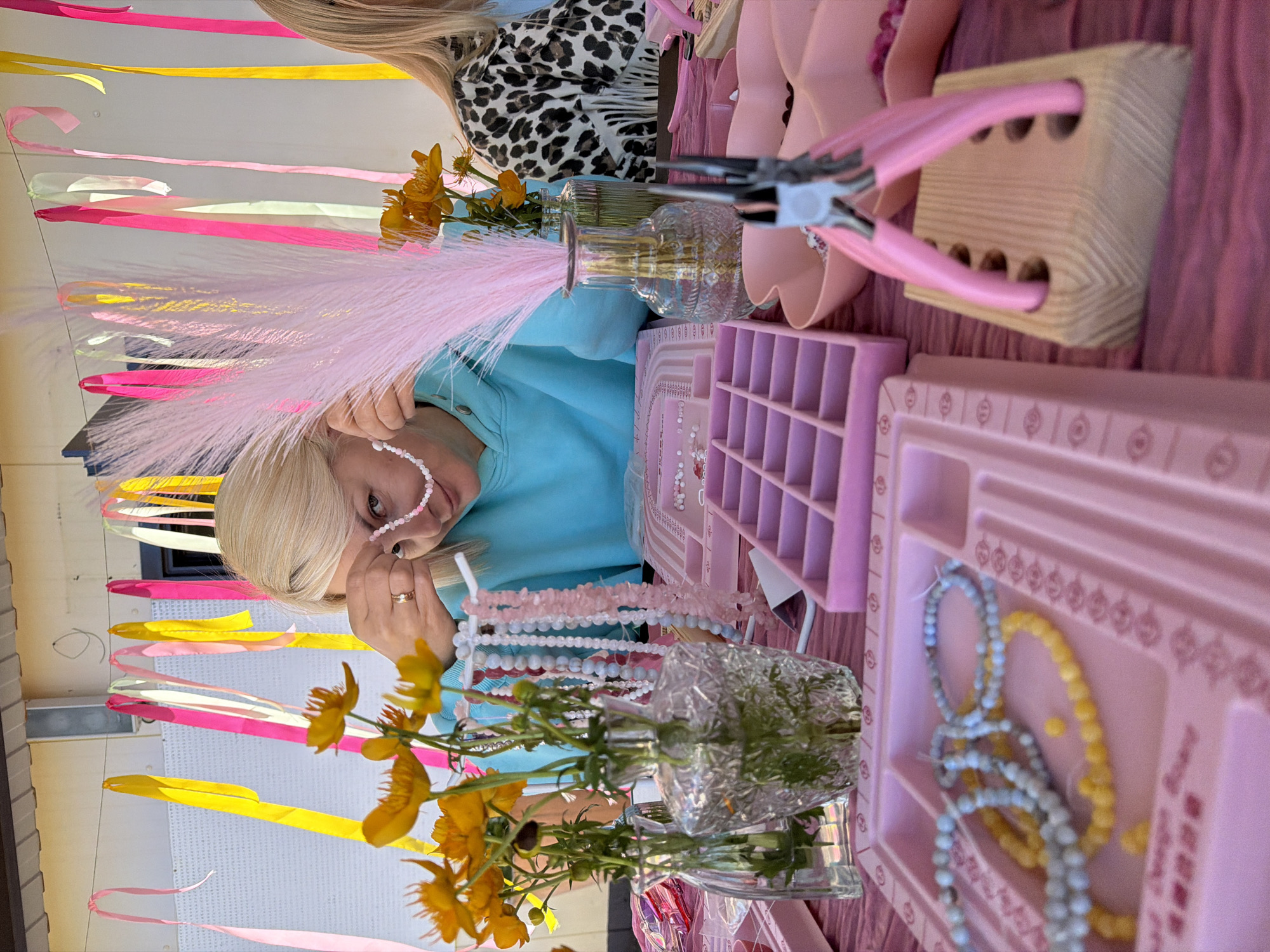 An outdoor scene with a table set up for jewelry making. A woman with blonde hair, wearing a light blue hoodie, is in the process of stringing beads onto a necklace. She is wearing a ring on her left hand. To her right, another woman with blonde hair, wearing a black and white leopard print top, is partially visible. The table is covered with a pink tablecloth and features various pink jewelry-making trays, bead organizers, vases with yellow flowers, and strands of colorful beads. Pink and yellow ribbons are strung up in the background. The environment suggests a creative or crafting event.