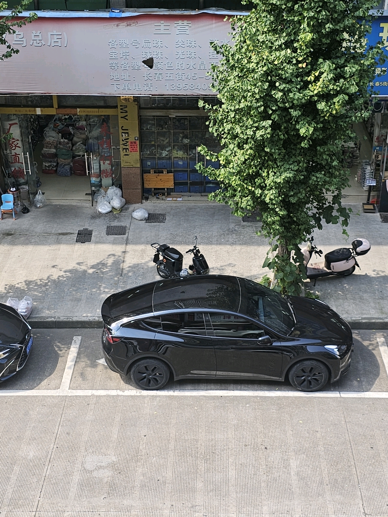 A street view in Yiwu, China, shows a black Tesla parked in a designated spot. To the right of the Tesla, a beige electric scooter is parked next to a tree. Further to the right, another black electric scooter is parked. In the background, a store with large windows displays various items, including what appears to be textiles or bags. Above the store, a white banner with Chinese text is visible. The street is paved with concrete, and shadows from a tree fall across the scene. The image is captured from an elevated perspective.