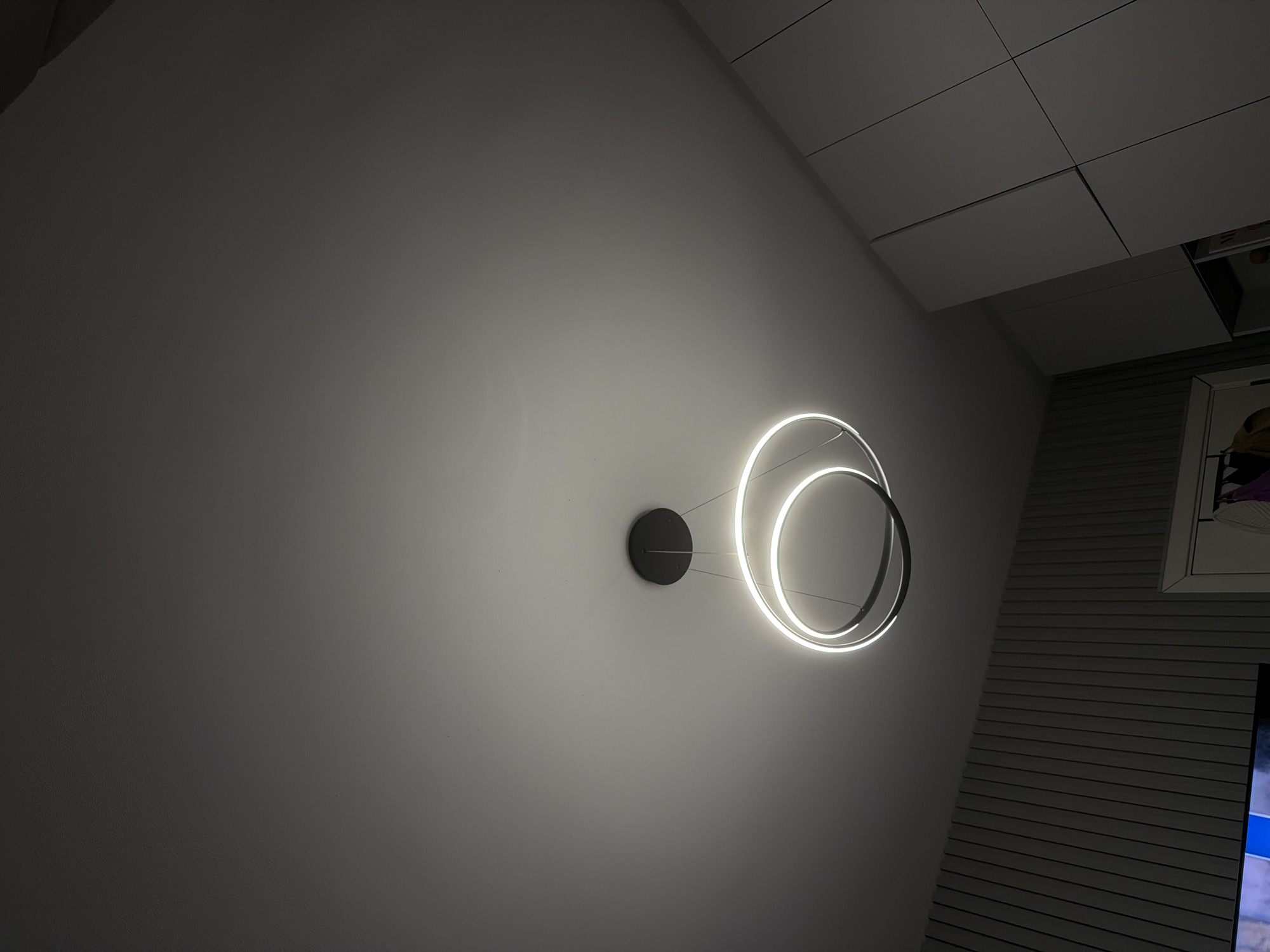 The image shows a modern, circular LED ceiling light fixture hanging from a white ceiling. The fixture consists of two concentric rings, one white and one black, with the white ring illuminated. Below the light, on the left, is a wall with vertical grey paneling. On the right, there are white closet doors. The scene appears to be an interior shot, likely in a residential setting in Ruse, Bulgaria. There are no people visible in the image.