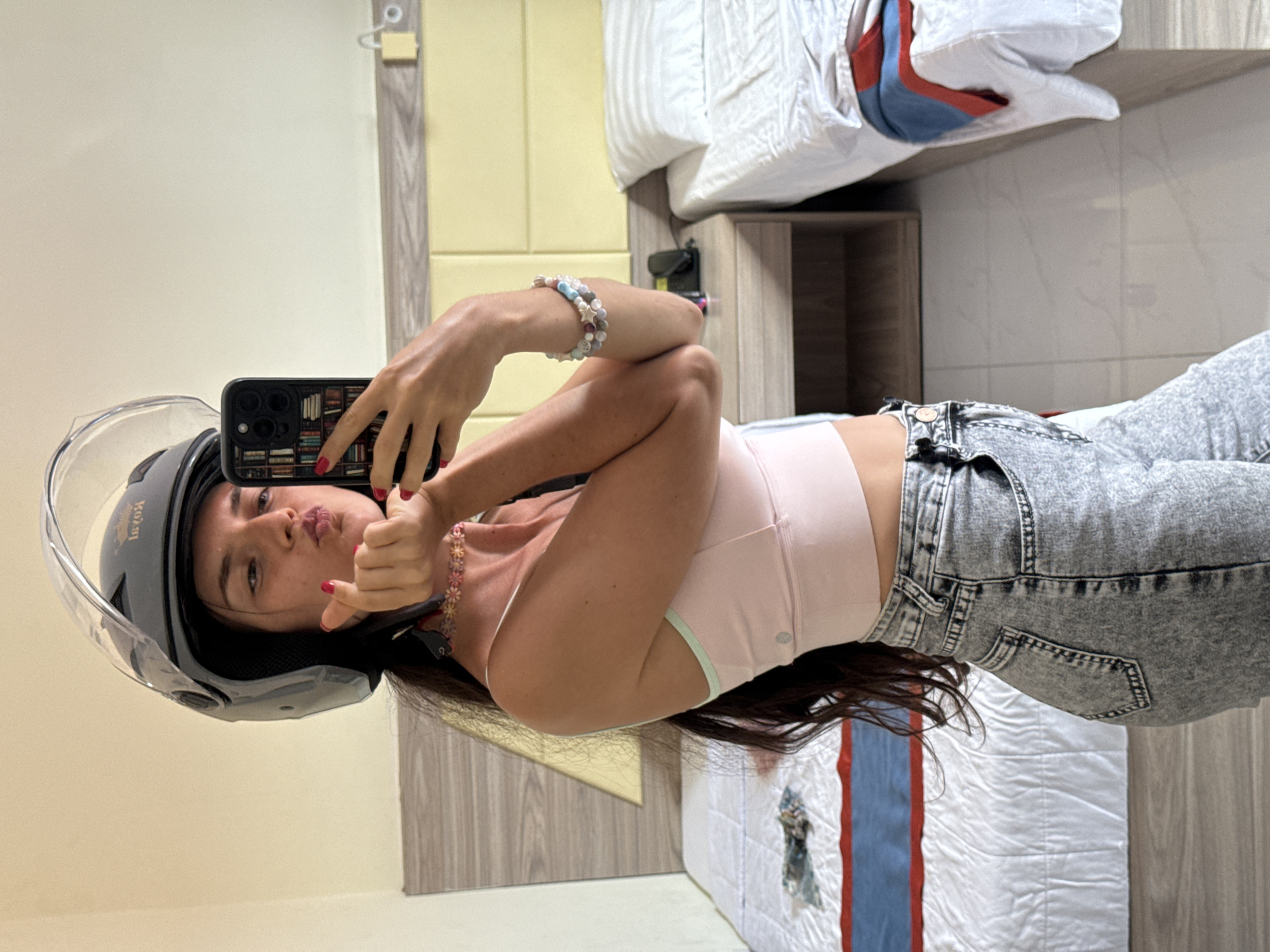 A young woman with long, dark hair is taking a mirror selfie in what appears to be a hotel room in Nha Trang, Vietnam. She is wearing a grey open-face helmet and a light pink crop top, paired with acid-wash denim jeans. Her left hand is holding her phone, and her right hand is touching her cheek, forming a heart shape with her fingers. She has bright red nail polish on. A colorful beaded bracelet is visible on her right wrist, and a floral choker necklace adorns her neck.
The room is furnished with two beds, each covered in white linens with a red and blue striped blanket. One bed is positioned against a wall with a wooden headboard and a yellow padded headboard above it. A bedside table with a phone and a lamp is situated between the two beds. The walls are a light cream color, and the floor is tiled. The overall impression is of a casual, possibly tourist-oriented accommodation. The image appears to have been taken during daylight hours.