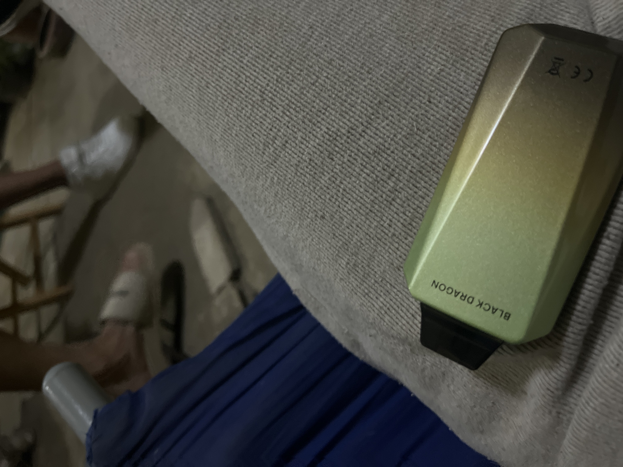 The image shows a close-up view of a person's lap, covered by grey fabric trousers. A light green and gold vape device with the text "BLACK DRAGON" and a CE marking is resting on the trousers. In the background, out of focus, are the legs of two individuals. One person is wearing white sneakers, and the other is wearing white slides. A blue object, possibly part of a chair or furniture, is visible on the left side of the frame. The scene appears to be outdoors, possibly on a patio or in a yard, with a concrete or dirt ground visible. The image does not provide any information about the location being Santa Ana, Venezuela.