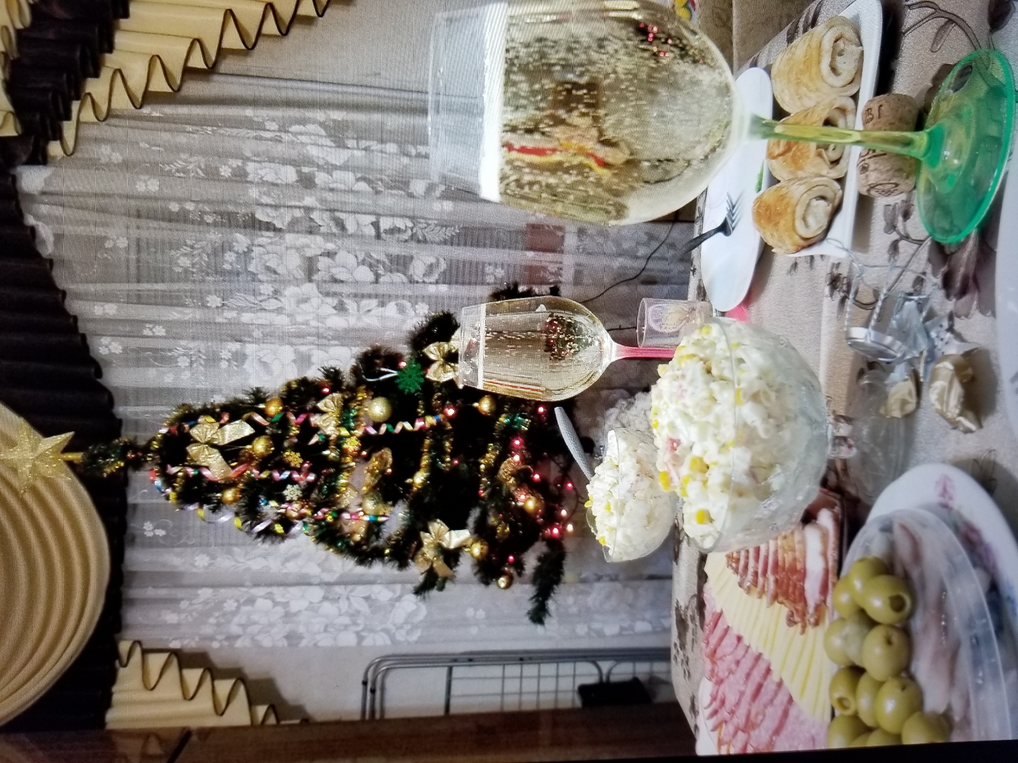 The image depicts a festive indoor scene, likely a holiday celebration, with a Christmas tree decorated with ornaments and lights as the focal point. The tree is positioned in front of sheer curtains. In the foreground, a table is laden with food and drinks. There are two glasses of champagne, several bowls of salad, a platter of sliced meats and cheese, a plate of olives, and rolled appetizers. Two wine corks and scattered confetti suggest a recent or ongoing celebration. The overall atmosphere is warm and inviting, indicative of a special occasion. The scene is consistent with a domestic setting, potentially in Tiraspol, Moldova, given the context provided. No people are visible in the image.