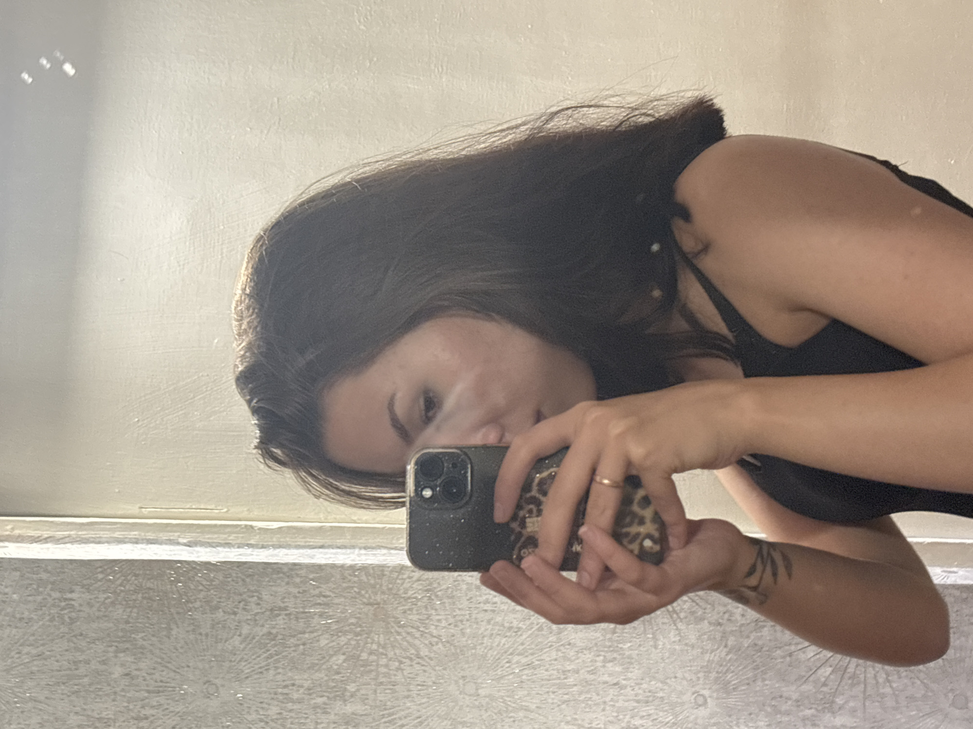 In the city of Inskoy, Russia, a young woman is captured in a mirror selfie, holding a smartphone adorned with a leopard-print case. She is wearing a black tank top, and her dark hair is styled with loose waves. Her left arm, visible in the frame, features a tattoo of a leafy branch. The background shows a wall with a textured, patterned wallpaper and a plain, cream-colored wall. The lighting suggests an indoor setting, possibly a bathroom or dressing area.