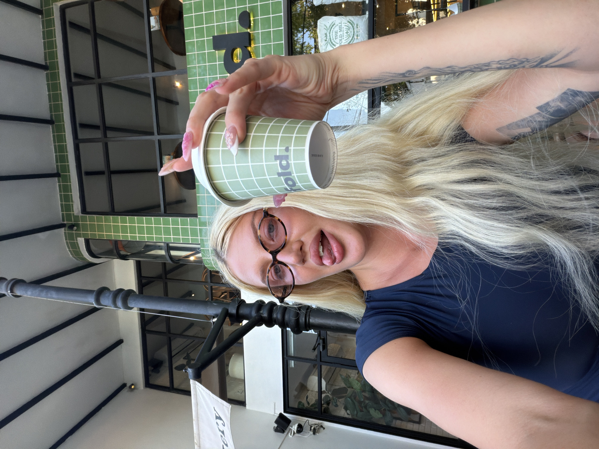 A young Caucasian woman with long blonde hair and a dark blue top is holding a green and white checkered coffee cup with the logo "fold." She is wearing oval-shaped glasses with a tortoiseshell pattern and sticking out her tongue. She has long, pink acrylic nails. The background shows a building with green tiled walls and large windows. There is a black lamppost in the foreground and a white awning to the left. The scene appears to be an outdoor seating area of a cafe or restaurant in North Kuta, Indonesia, based on the provided context.