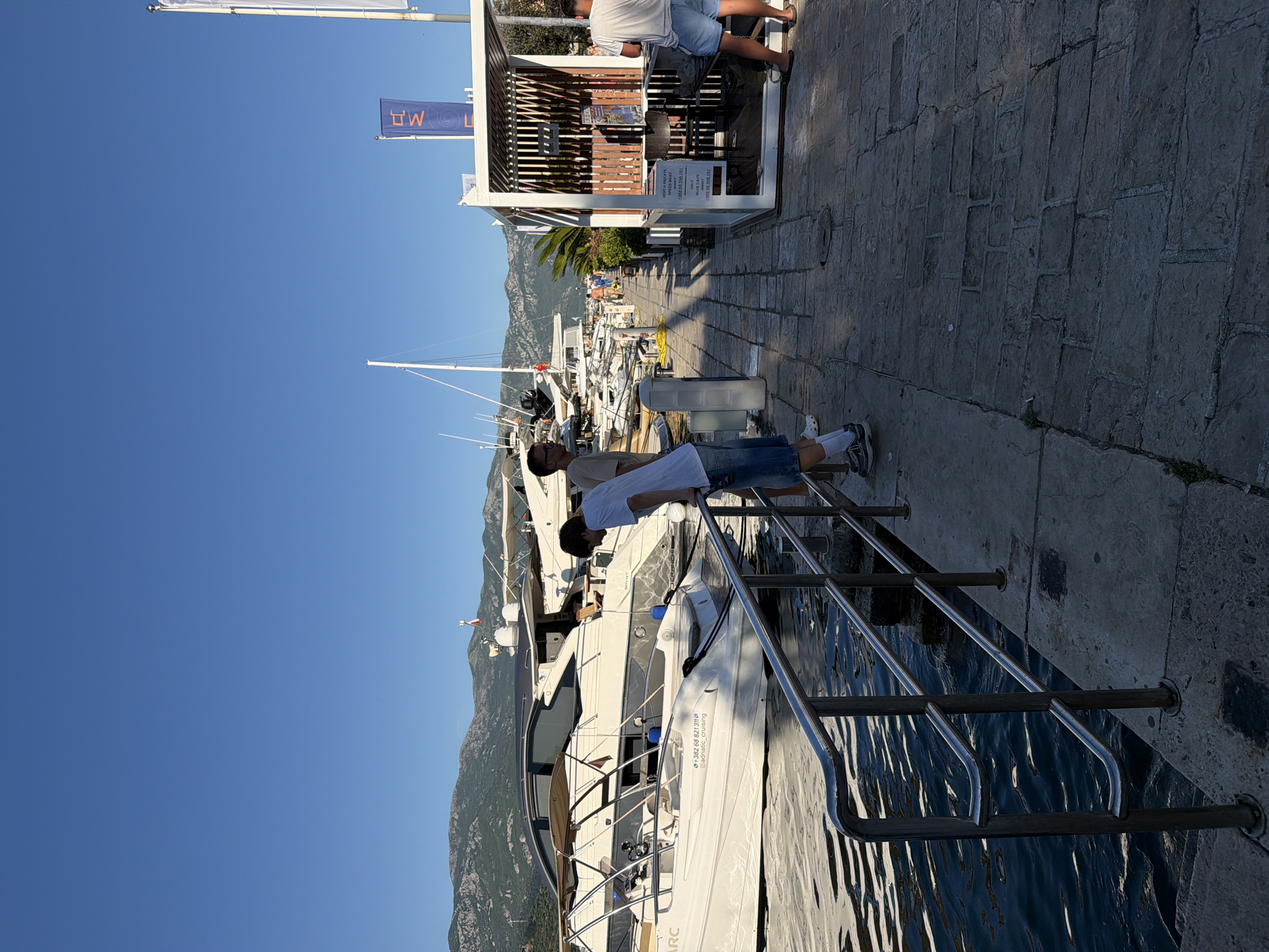 In Budva, Montenegro, a waterfront promenade is captured under a clear blue sky. The scene is dominated by several luxury yachts moored in the harbor, their sleek designs reflecting in the calm, dark water. A stone walkway, bordered by a polished metal railing, lines the edge of the water. Two young men are visible on the walkway, one leaning over the railing and gazing at the boats, while the other stands a few feet behind him. Further down the walkway, a white open-air structure with a slatted roof serves as a small kiosk, where a sign advertises boat rentals. Behind the kiosk, the distant green slopes of mountains rise, adding a natural backdrop to the urban marina. The overall atmosphere is one of leisure and sophistication, typical of a Mediterranean coastal town.