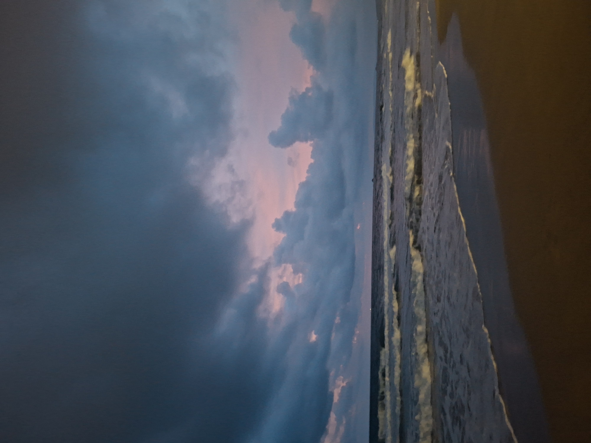 The image shows a beach scene at dusk on Phu Quoc Island, Vietnam. The sky is filled with dramatic, dark clouds tinged with pink from the setting sun. Waves are gently breaking on the shore, leaving a foamy white line on the wet sand. The beach itself is dark and appears to be empty. In the distance, the sea stretches to the horizon, merging with the dusky sky. The overall mood is serene and tranquil, despite the foreboding clouds.