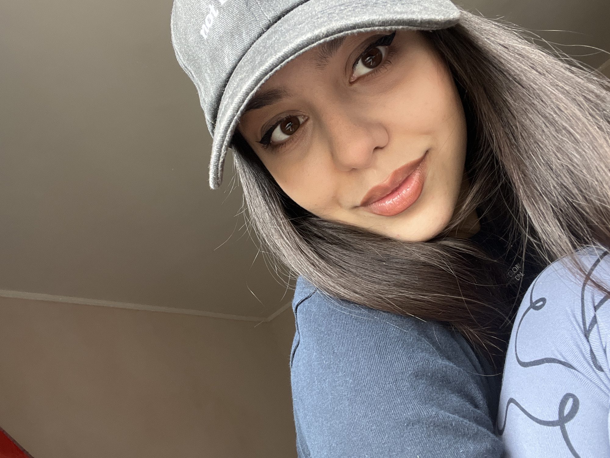 A close-up, angled portrait depicts a single female subject in what is reported to be the city of Zheglartsi, Bulgaria. The subject, a young adult, looks directly towards the camera with a subtle smile. She has dark brown, straight hair and brown eyes, enhanced with black winged eyeliner and a light red lip product. She wears a grey denim-textured baseball cap, with partial white text visible on the front. Her attire includes a dark blue long-sleeved garment, partially covering another dark garment with fragmented white text, and a light blue garment with black wavy lines, visible on her lower left. The background consists of an out-of-focus, plain light brown wall, with a faint vertical lighter line visible towards the upper right.