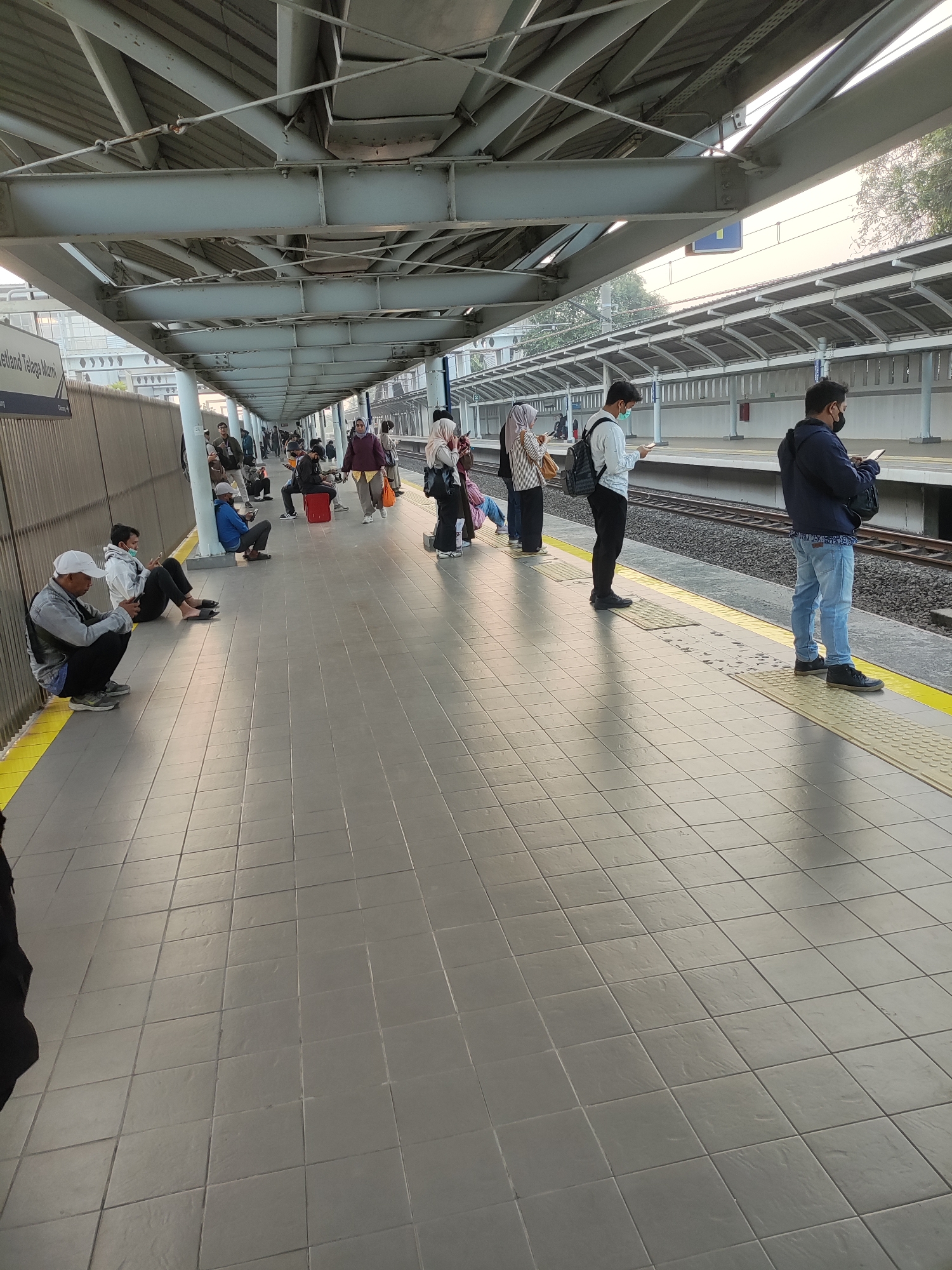 The image depicts a train station platform in Telagamurni, Indonesia, with several individuals waiting. The platform is tiled with a gray, square pattern and bordered by a yellow safety line. A covered walkway with a metal roof structure supported by beams extends along the platform.
In the foreground, on the left, a person wearing a white cap and a denim vest over a light-colored hoodie is crouched on the ground, looking down. Next to them, further back and seated on the platform, is another individual in a white hoodie and dark pants, also looking at their phone.
Further down the platform, a group of people are standing and sitting. Some are looking at their phones, while others are engaged in conversation. One person, wearing a blue jacket, stands by the tracks, looking at their phone. Another person, in a dark jacket and jeans, is also standing and looking at their phone. Several women are visible, some wearing headscarves. One woman is holding a red suitcase.
The environment appears to be an outdoor urban setting, with the railway tracks running alongside the platform. The lighting suggests it might be late afternoon or early morning, with a soft, diffused light. The background shows the station's infrastructure, including a fence and the continuation of the covered walkway. The overall scene is one of quiet waiting, with people occupied with their personal devices.
