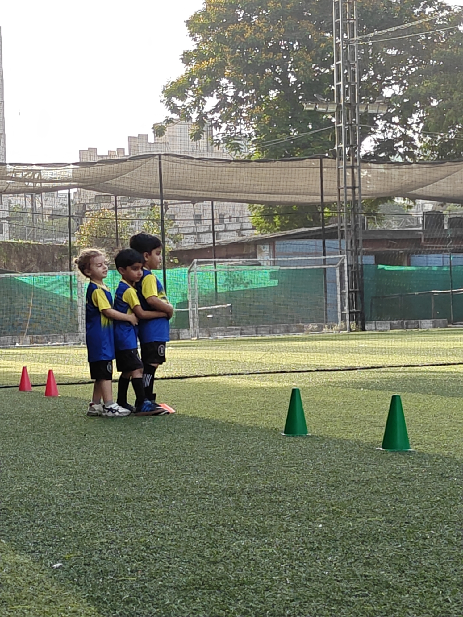 Three children in matching blue and yellow jerseys stand together on a green turf field. Orange cones are visible to their left, and green cones are to their right. In the background, there's a structure covered with netting, a football goal, trees, and what appears to be buildings under construction. The scene appears to be outdoors during daylight. Given the context, it's likely the location is a sports field in Mumbai, India.