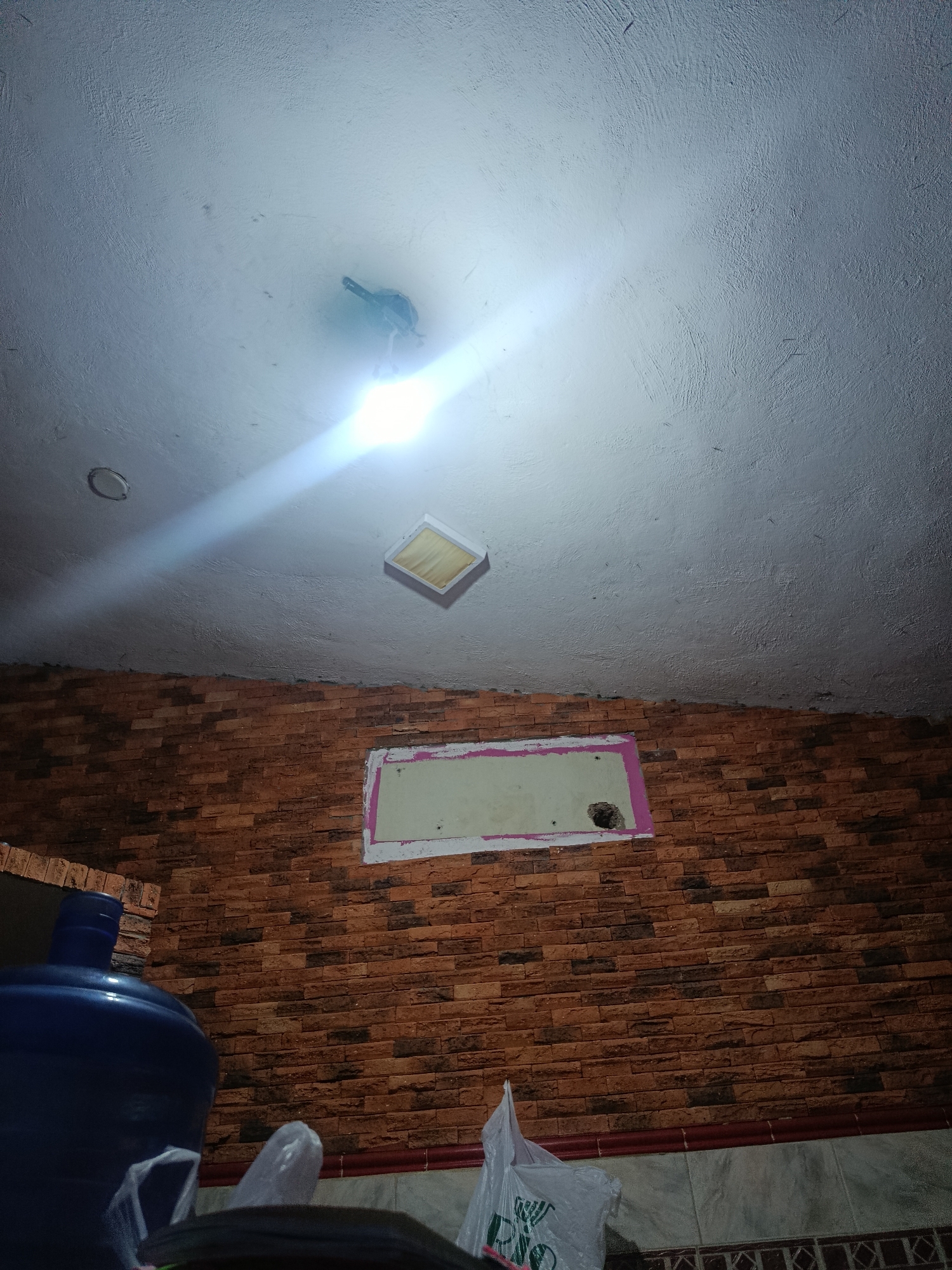 The image depicts an indoor scene with a textured orange brick wall and a white ceiling. On the ceiling, there is a light fixture that is emitting a bright glare, and another square light fixture is partially visible. Below the ceiling, a rectangular opening with a pink border is present on the brick wall, with a hole in the center of it. To the left of the frame, a large blue water jug is visible, along with plastic bags and other objects on a surface. The lighting suggests it is likely nighttime or the room is dimly lit, with the ceiling light being the primary light source. The overall impression is that of a utility or storage area.