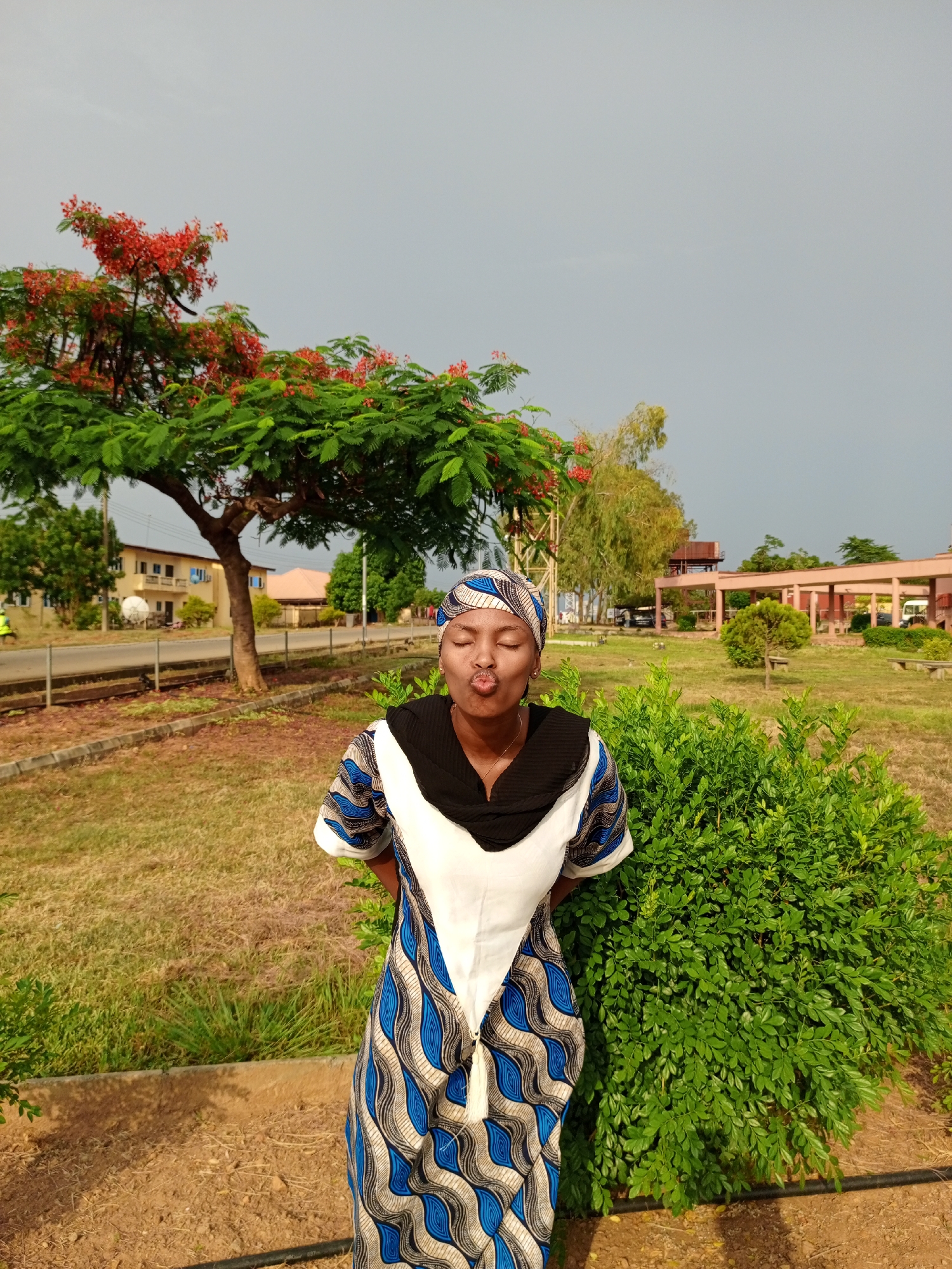 The image shows a woman standing in a grassy area with buildings and trees in the background. She is wearing a colorful patterned dress and a headscarf, and she is making a kissing face. Behind her is a green bush, and a tree with red flowers is visible on the left side of the frame. In the background, there is a building with a red roof and a covered walkway. The sky is overcast. The scene appears to be in Keffi, Nigeria.
