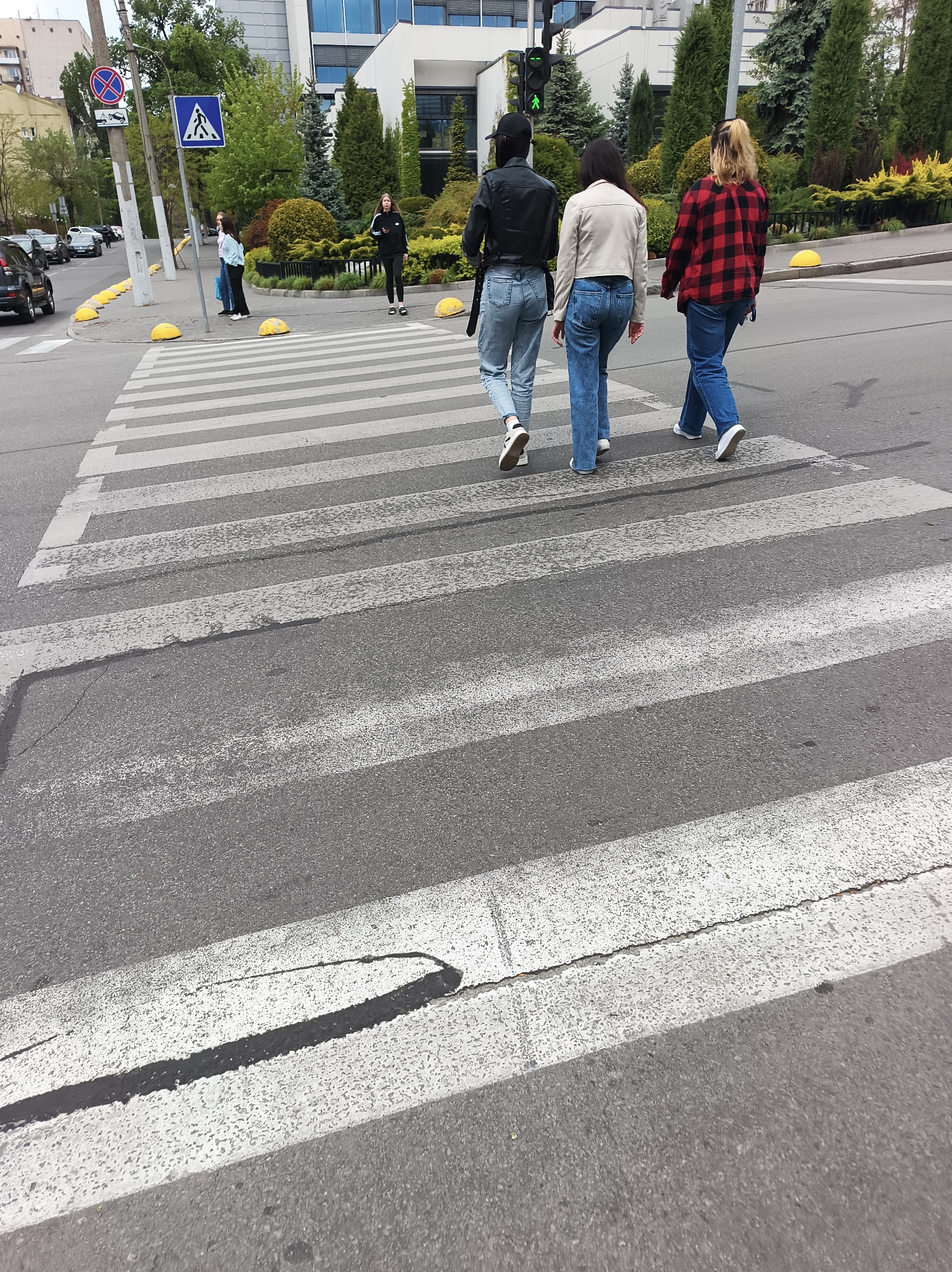The image shows three women crossing a pedestrian crosswalk in Kyiv, Ukraine. The women are wearing casual clothing, including jeans, jackets, and sneakers. They are walking from left to right in the frame. A pedestrian crossing sign is visible on the left side of the road. In the background, there are buildings, trees, and other pedestrians. A row of yellow road bollards runs along the edge of the sidewalk. A car is driving on the street to the left. The crosswalk itself is worn, with visible cracks in the asphalt.