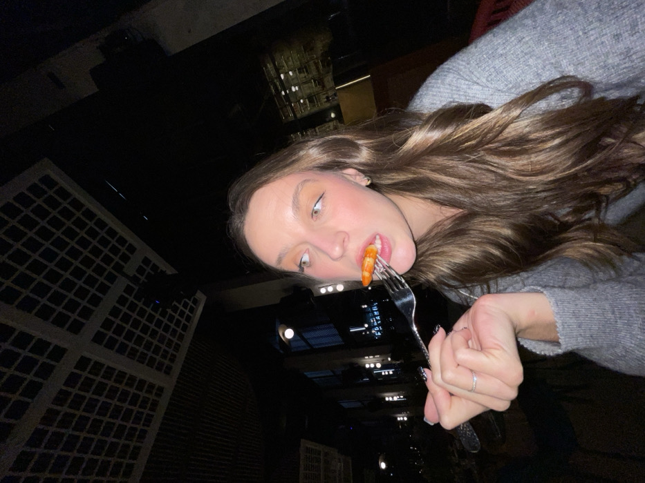 A young woman with long brown hair eats a shrimp on a fork. She wears a gray sweater. The backdrop is a dark night scene with buildings visible, illuminated by lights. Her manicure is visible, with an intricate black design. She is fair-skinned with pink blush on her cheeks. The perspective is unusual, tilted and looking up at her face.