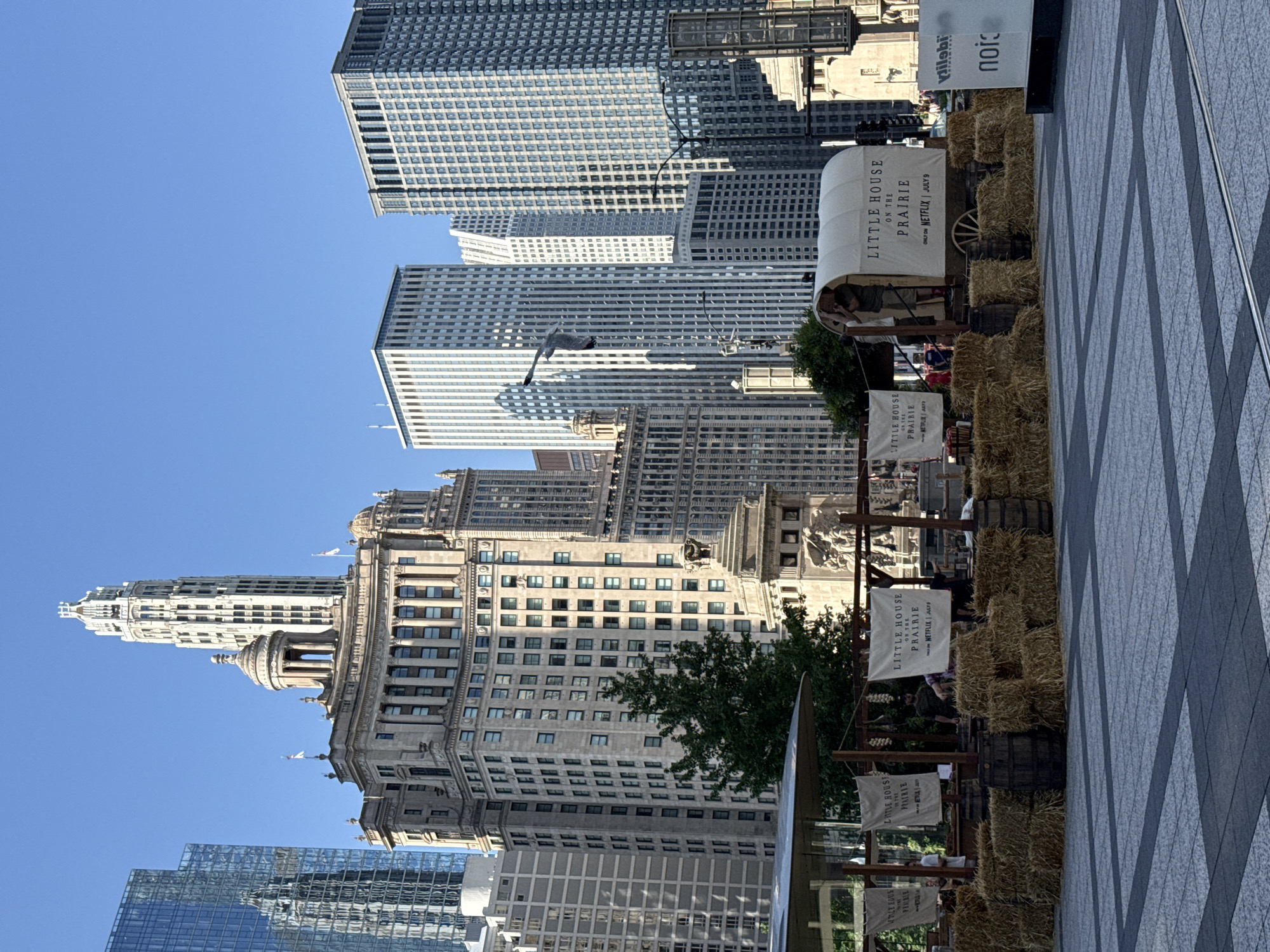 The image depicts a street scene in Chicago, United States of America, featuring a promotional display for "Little House on the Prairie." In the foreground, a covered wagon, reminiscent of those used in the American West, is set up on a paved plaza. The wagon is adorned with banners that read "LITTLE HOUSE ON THE PRAIRIE" and "ONLY ON NETFLIX | JULY 9." The display is surrounded by hay bales and wooden barrels. Several people are visible interacting with the display, some standing near the wagon and others behind it.
Behind the promotional setup, the cityscape of Chicago is visible, characterized by numerous skyscrapers of varying architectural styles. Notable buildings include a tall, art deco skyscraper with a distinctive spire and a modern, glass-faceted building reflecting the sky. The background is dominated by a clear blue sky. A green tree stands to the left of a monument. A seagull is captured in flight in the mid-ground. The overall scene appears to be set during daylight hours.