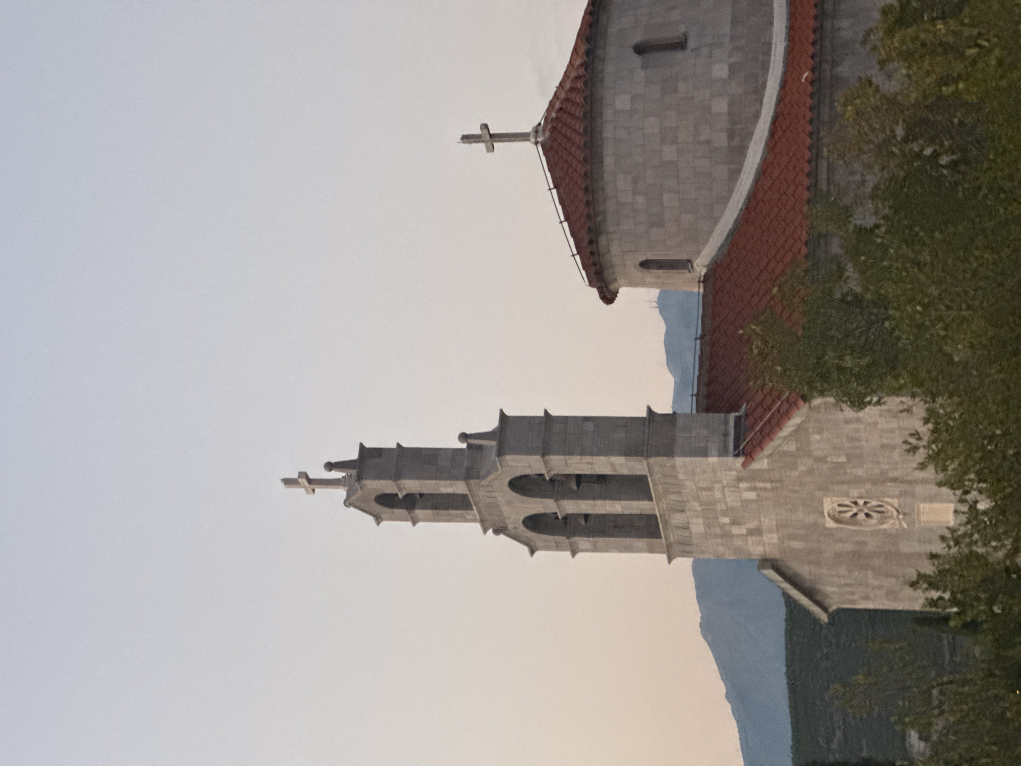 This is a vertical, eye-level, outdoor, daytime shot of the Church of Saint Catherine in Kovačko Polje, Montenegro, taken from a slightly elevated position. The church features a bell tower to the left and a domed structure to the right, both topped with crosses. The bell tower has three archways and is constructed of stone blocks. The domed section also appears to be stone, with a red tiled roof. In the foreground, there are dark green trees, and in the background, blue mountains are visible against a pale, hazy sky. The image has a grainy texture.