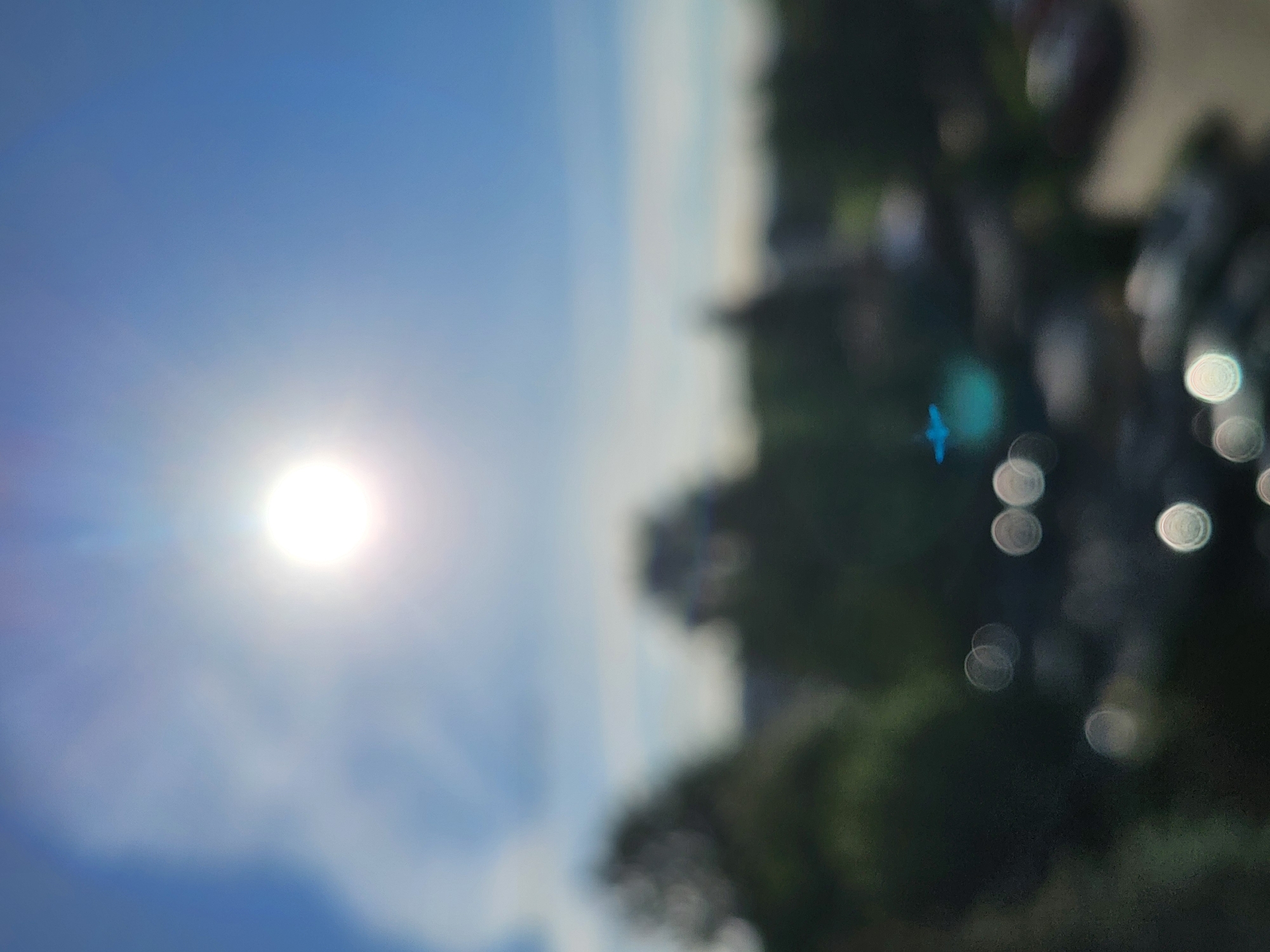 The image shows a blurry outdoor scene under a bright sun in a blue sky with scattered clouds. There's indistinct greenery suggestive of trees and possibly buildings on the right side, with some out-of-focus circular highlights. A small, bright blue streak appears near the center-right. The overall scene lacks specific details to determine a precise location.