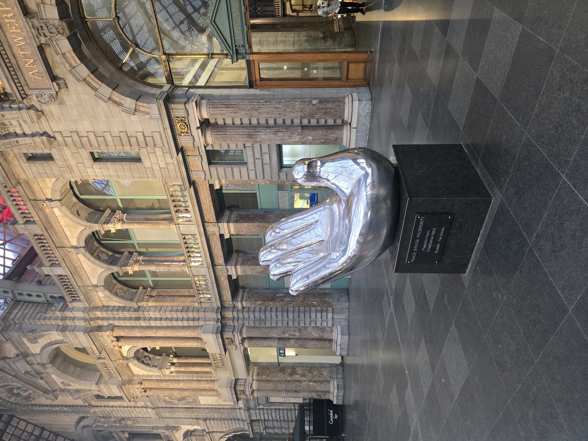 The image depicts the interior of Antwerp Central Station, also known as the 'Iron Cathedral', in Antwerp, Belgium. The grand architecture of the station is visible, with its ornate stone facade, tall columns, arched windows, and a glass-domed ceiling not fully in view. In the foreground, a large, polished silver sculpture of an open hand with its palm facing upwards rests on a dark granite pedestal. The inscription on the pedestal reads: "PEACE BRINGS PROSPERITY Peaceful Hand Vreedzame Hand Sculptor: Yan Shulten". The floor is tiled with dark grey granite squares. To the right, a person in a light-colored shirt and dark pants is walking away from the camera, near an open wooden doorway. To the left, a black canopy tent with "Corsendonk" written on it is visible. The lighting suggests it is daytime, with sunlight filtering through the station's structure, creating reflections on the floor and the sculpture.