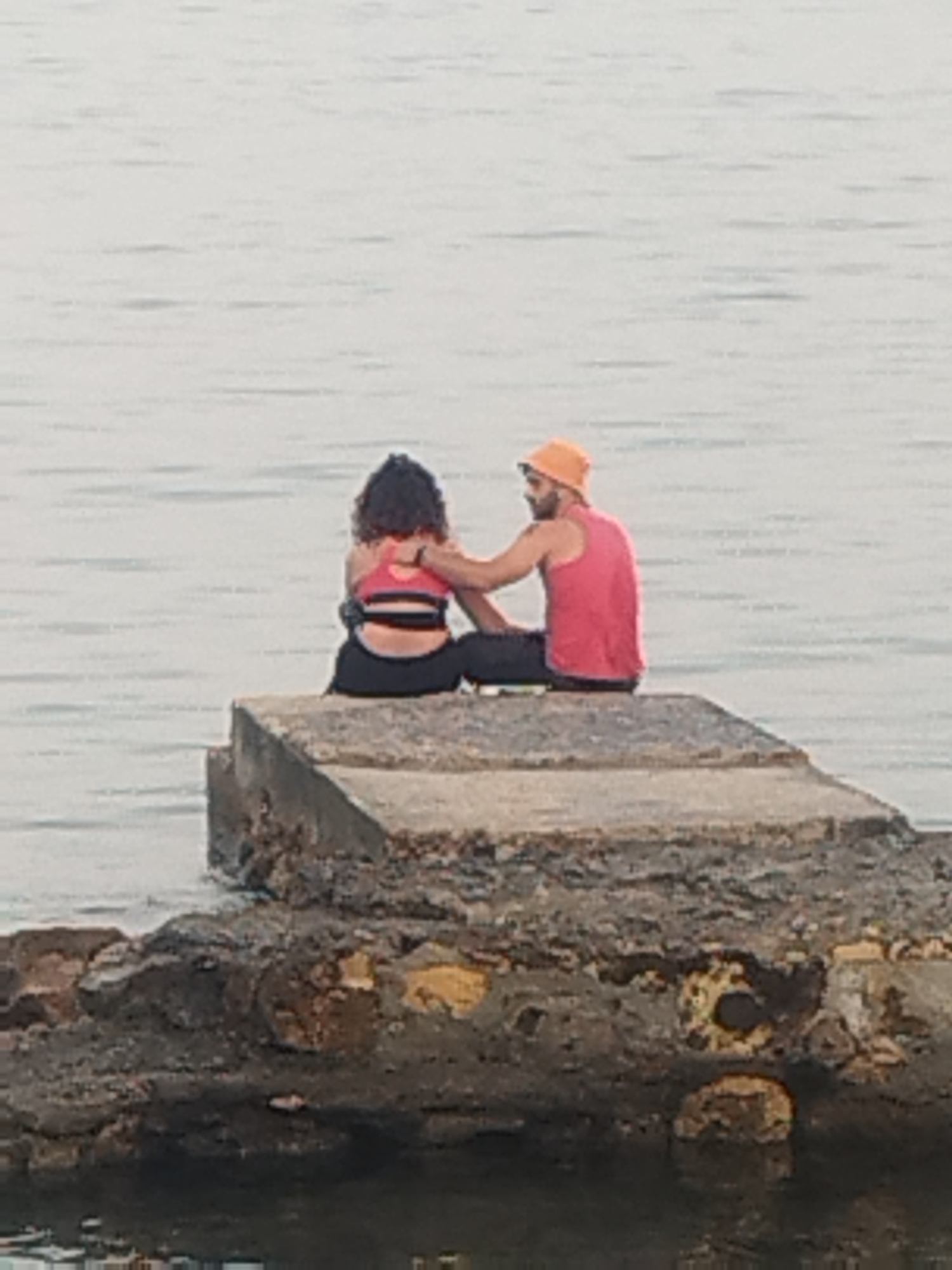 Two individuals, one with dark curly hair wearing a pink sports bra and black leggings, and another with dark hair and a beard wearing a pink tank top and an orange bucket hat, are seated on a concrete structure at the edge of the water. The person with the hat has their arm around the other person's shoulder. The concrete structure appears weathered and sits on top of a rocky embankment. The background is dominated by calm, pale water. The location is the Port Vauban in Antibes, France.
