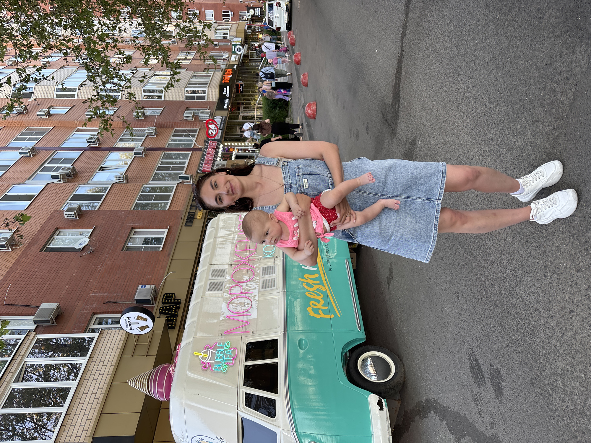 A woman is holding a baby on a street in Krasnodar, Russia. The woman, standing in front of a vintage-style ice cream truck, is wearing a denim dress and white sneakers. The baby is dressed in a pink outfit. Behind them is a street with pedestrians and a multi-story brick building with various signs, including one for "24". The ice cream truck has lettering in Russian and English ("Fresh" and "Bubble Tea"), with an ice cream cone decoration on top. The street is asphalt.
