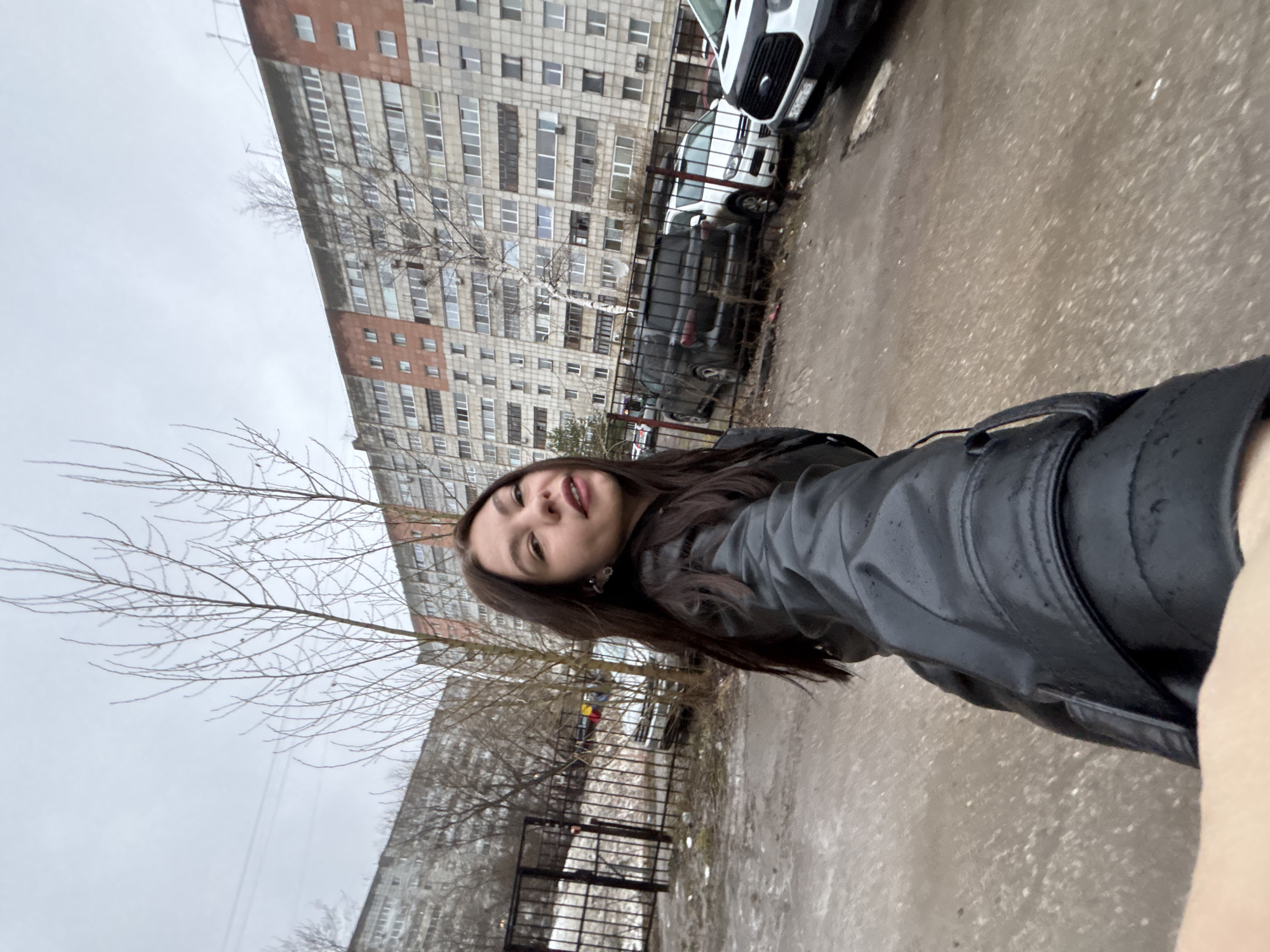 A waist-up selfie of a young woman wearing a black leather jacket. She is looking at the camera with a neutral expression. Behind her is a large, grey apartment building with many windows. There are bare trees in front of the building. Several cars are parked along the side of the road, and a metal fence is visible in the foreground. The ground is wet and appears to be concrete or pavement. The photograph was taken in Perm, Russia.