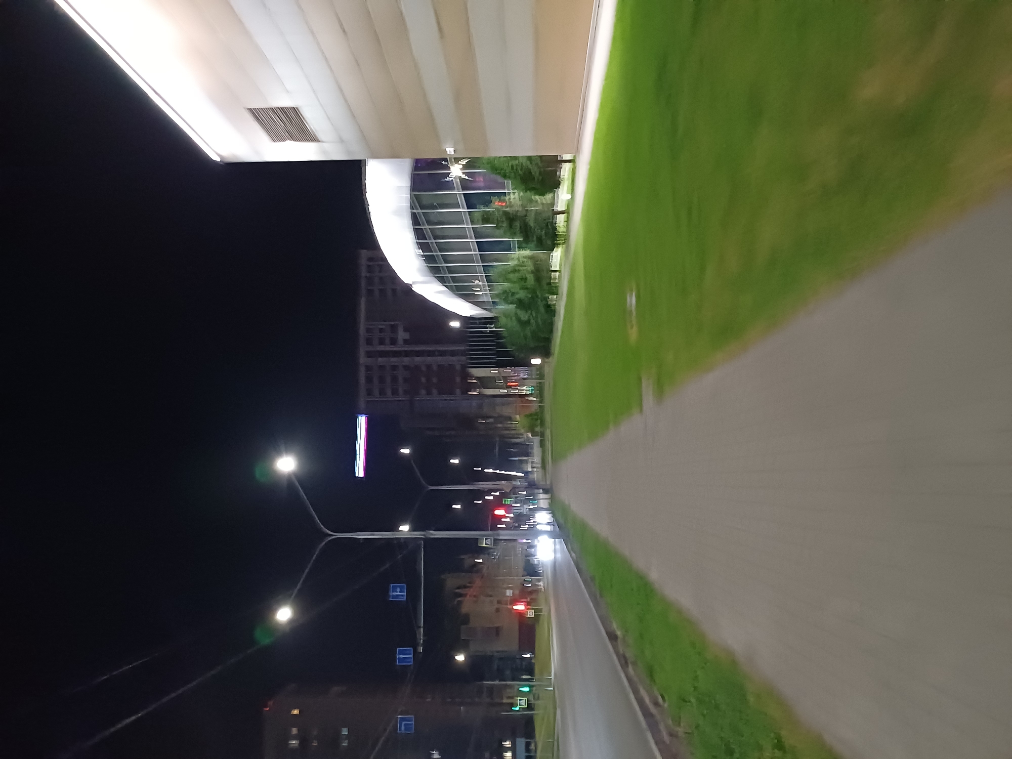 The image shows a nighttime scene in a city. On the left, there is a street with cars and streetlights. A pedestrian walkway runs alongside the street, bordered by a grassy area. In the background, there are buildings of various heights, some illuminated, and a large modern building with a curved glass facade. The sky is dark, indicating it is night. The image is taken from a low angle, possibly from a moving vehicle or bicycle, as evidenced by the motion blur in the foreground.