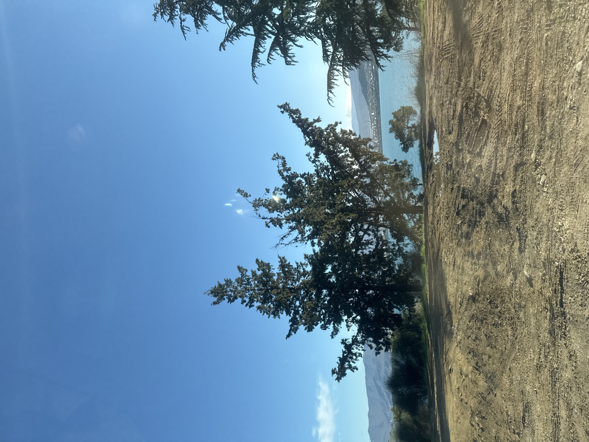 The image displays a landscape featuring a body of water, possibly a lake, bordered by a dirt path and foliage. The sky is clear blue, occupying a large portion of the frame. Trees are present in the foreground and midground, framing the scene. Distant mountains are visible across the water. The dirt path shows tire tracks and appears recently disturbed. The photograph appears to be taken at ground level, slightly angled upwards. There is no evidence to support the claim that the photograph was taken in Tbilisi, Georgia.
