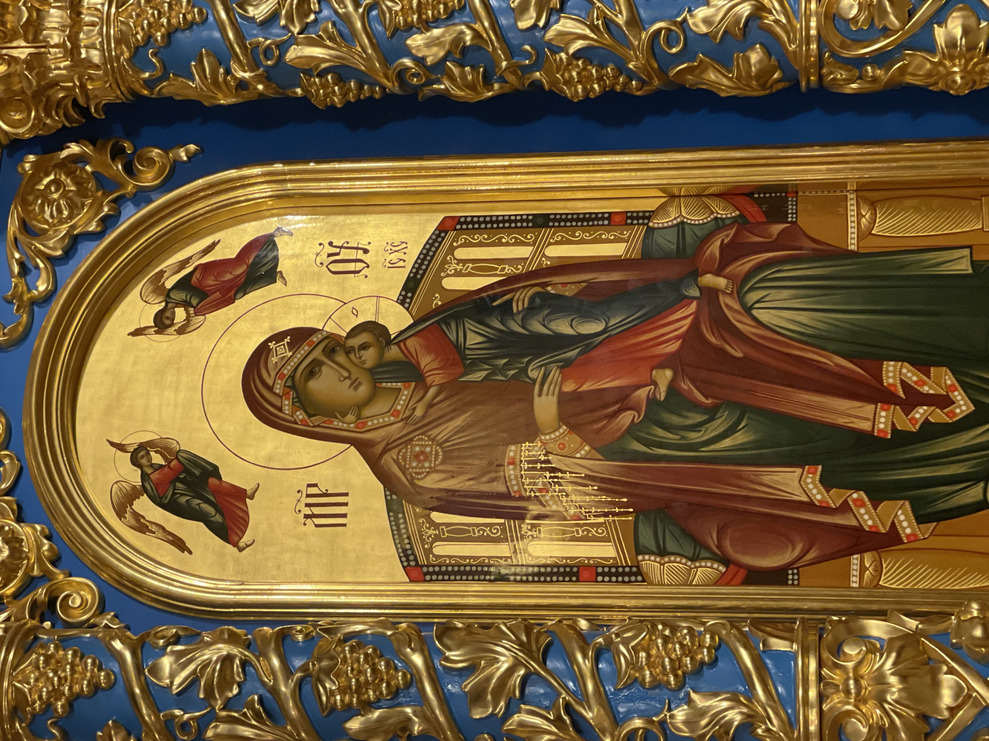 The image shows an Orthodox icon of the Virgin Mary holding the infant Jesus. Two angels are depicted on either side of them. The icon is framed in a golden ornate border, surrounded by a blue and gold decorative background. The painting style is Byzantine. The location is likely in a church in Moscow, Russia, given the prevalence of Orthodox iconography in the city.