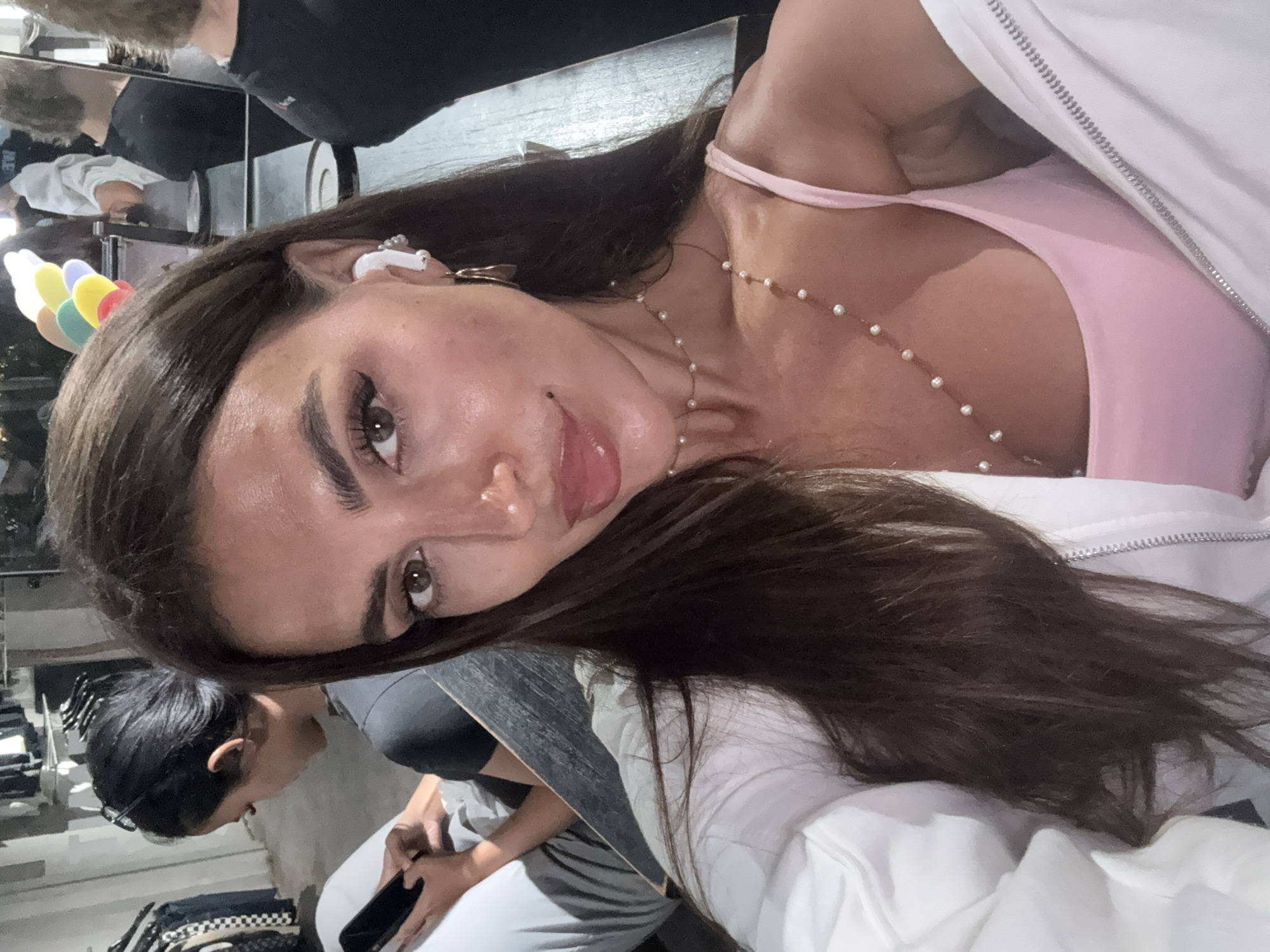 A woman with long, dark brown hair and light skin is posing for a selfie indoors. She's wearing a pink top, a white jacket, and a pearl necklace. An earbud is visible in her left ear.
In the background, other people are present, one of whom is using a phone. Various objects are visible, including a table, possibly a mirror, and items that suggest the location is in North Kuta, Indonesia.