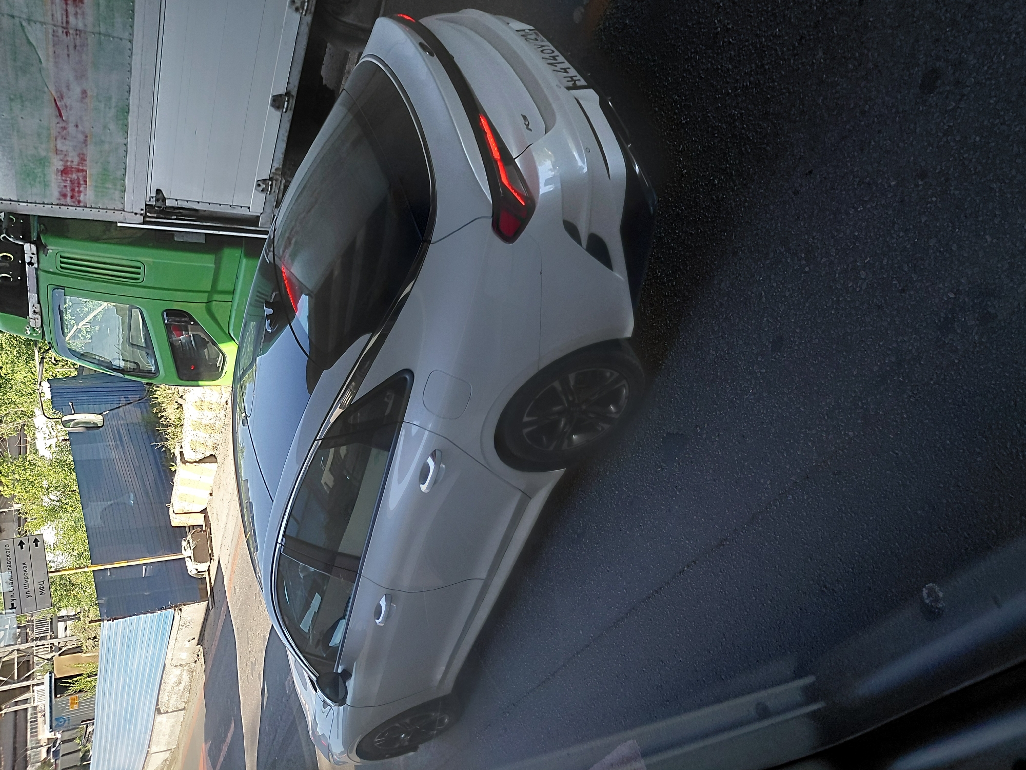 A white sedan is parked on an asphalt road next to a green truck. The sedan has a sleek design and black rims. The truck is a cargo truck with a white boxy trailer. In the background, there are construction barriers and signage indicating directions to "ул. Станиславского" and "ул. Широкая". The scene appears to be in an urban area under daylight.