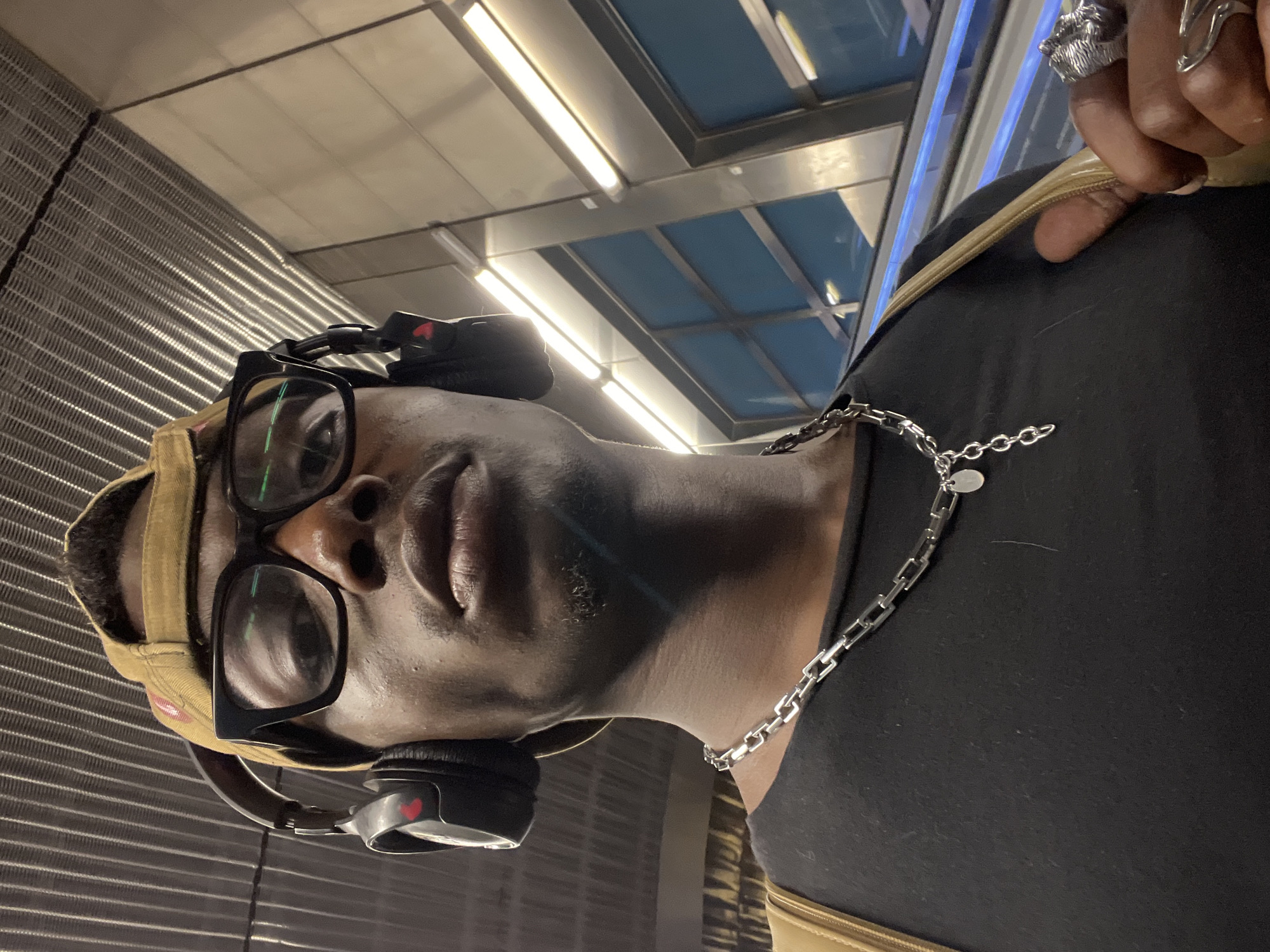 The image captures a close-up portrait of a person, likely a young adult male with dark skin, looking directly towards the camera. The person is wearing black-framed glasses and black over-ear headphones, which feature a small red heart symbol on one ear cup. A tan baseball cap is partially visible on their head. They are dressed in a plain black t-shirt, adorned with a silver chain necklace that has a small, round pendant. Several distinct rings are worn on the fingers of their right hand, which is visible near their shoulder.
The background suggests an indoor, modern public environment. To the person's left, a wall with a prominent horizontal corrugated or grated pattern extends upwards. Above and to the right, the ceiling features bright fluorescent strip lights set within metal frames, alongside sections of blue panelling. The artificial lighting illuminates the scene. The person's expression appears neutral or contemplative. The location context implies this could be a setting within Barcelona, Spain, such as a transit station or a contemporary building. No other discernible text is visible besides the heart icon on the headphones.