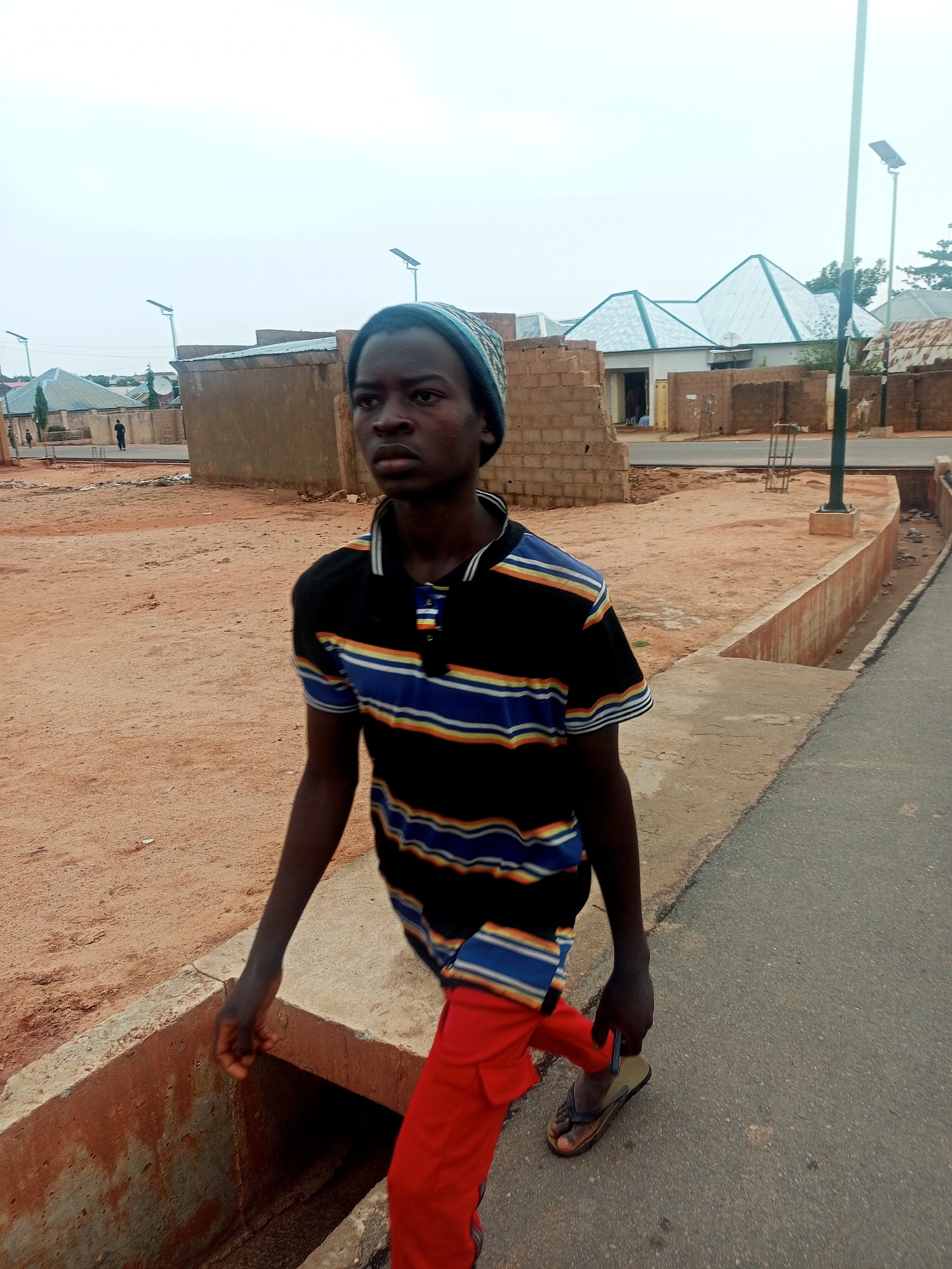 A young, dark-skinned male wearing a striped polo shirt with black, blue, and gold stripes, red trousers, and a knit cap is walking alongside a paved road in what appears to be a residential area. He is wearing flip-flops. To his left is a concrete drainage ditch. Behind him are various buildings constructed of unfinished brick and other materials. There are lampposts along the street. The sky is overcast. The location is Tempure, Nigeria.