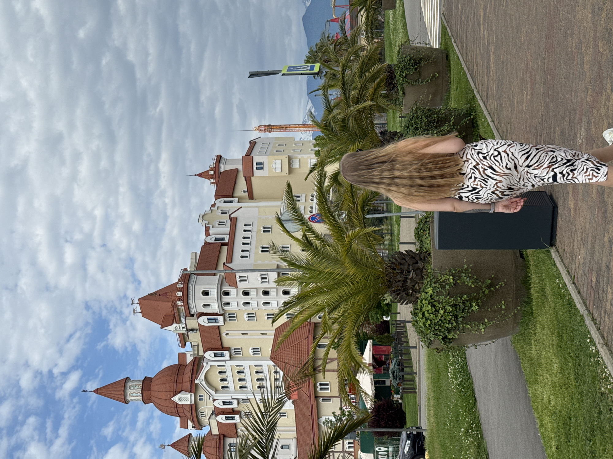 The image depicts a woman with long blonde hair walking along a paved sidewalk in Sirius, Russia. She is wearing a zebra-striped dress and white shoes. The sidewalk is lined with large concrete planters filled with palm trees and trailing vines. A grassy area lies between the sidewalk and a lower pathway.
Behind the woman and the palm trees is a large, ornate building with red roofs and beige walls, featuring numerous windows and turrets. A pedestrian crossing sign is visible above the palm trees. In the background, distant mountains can be seen under a cloudy sky. A tall crane is situated near the building. A black car is visible at the bottom left.