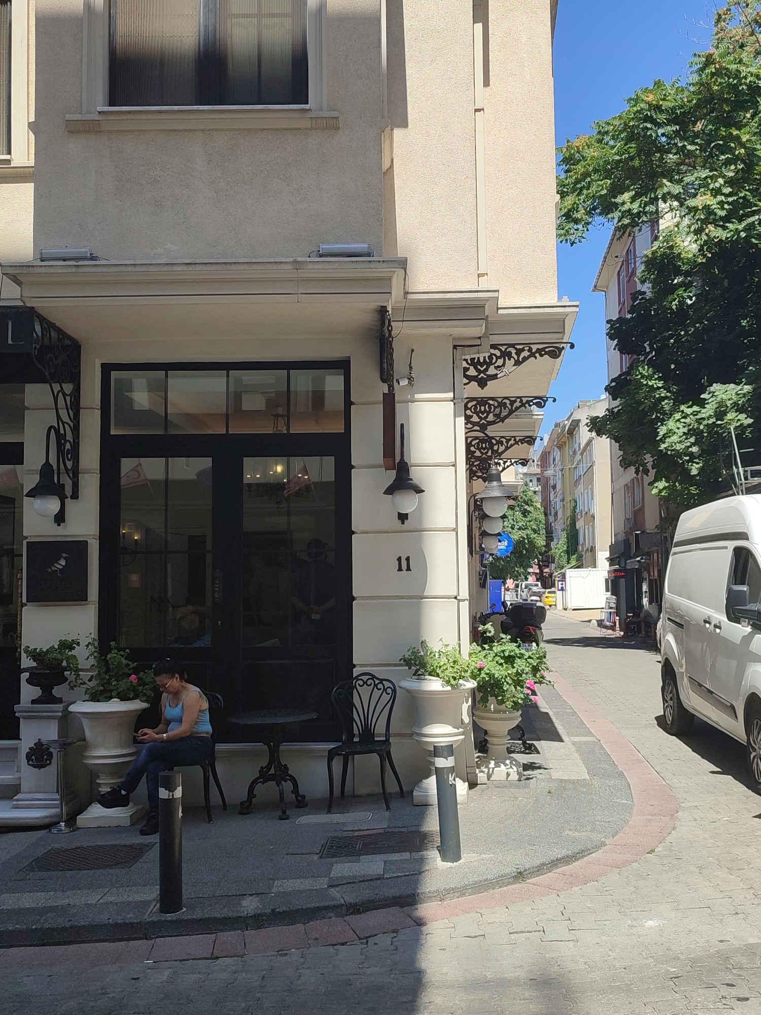 The image captures a street scene in Kadıköy, Turkey, on a sunny day. The foreground features a woman seated on a black wrought-iron chair outside a building. She is wearing a blue tank top and dark jeans and is looking at her phone. In front of her is a small, round, black wrought-iron table. Flanking the entrance to the building are two large white planters filled with green foliage and pink flowers. The building itself is cream-colored with a dark-framed glass door and windows above. Decorative black wrought-iron elements adorn the facade. To the right of the building, a white van is parked on the street, which is paved with cobblestones and has a pink-lined pedestrian walkway. In the background, a street lined with multi-story buildings and trees extends into the distance. The sky is clear and blue.