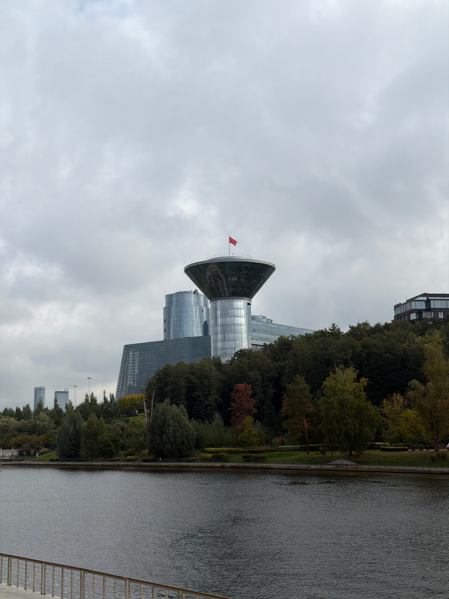 A wide shot shows a body of water in the foreground, with a metallic railing along the bank. Across the water, a park with dense trees stretches along a hillside. Beyond the park, several modern buildings rise into the sky, including a distinctive circular structure with a red flag on top. The sky is overcast with gray clouds, suggesting a cool, possibly autumn day. The buildings appear to be part of a business or administrative complex, with large glass facades reflecting the cloudy sky. In the distance, two taller, rectangular buildings are visible. The overall scene conveys a sense of urban development integrated with natural green spaces. The location is in Khimki, Russia, specifically depicting the area near the Moscow Canal.