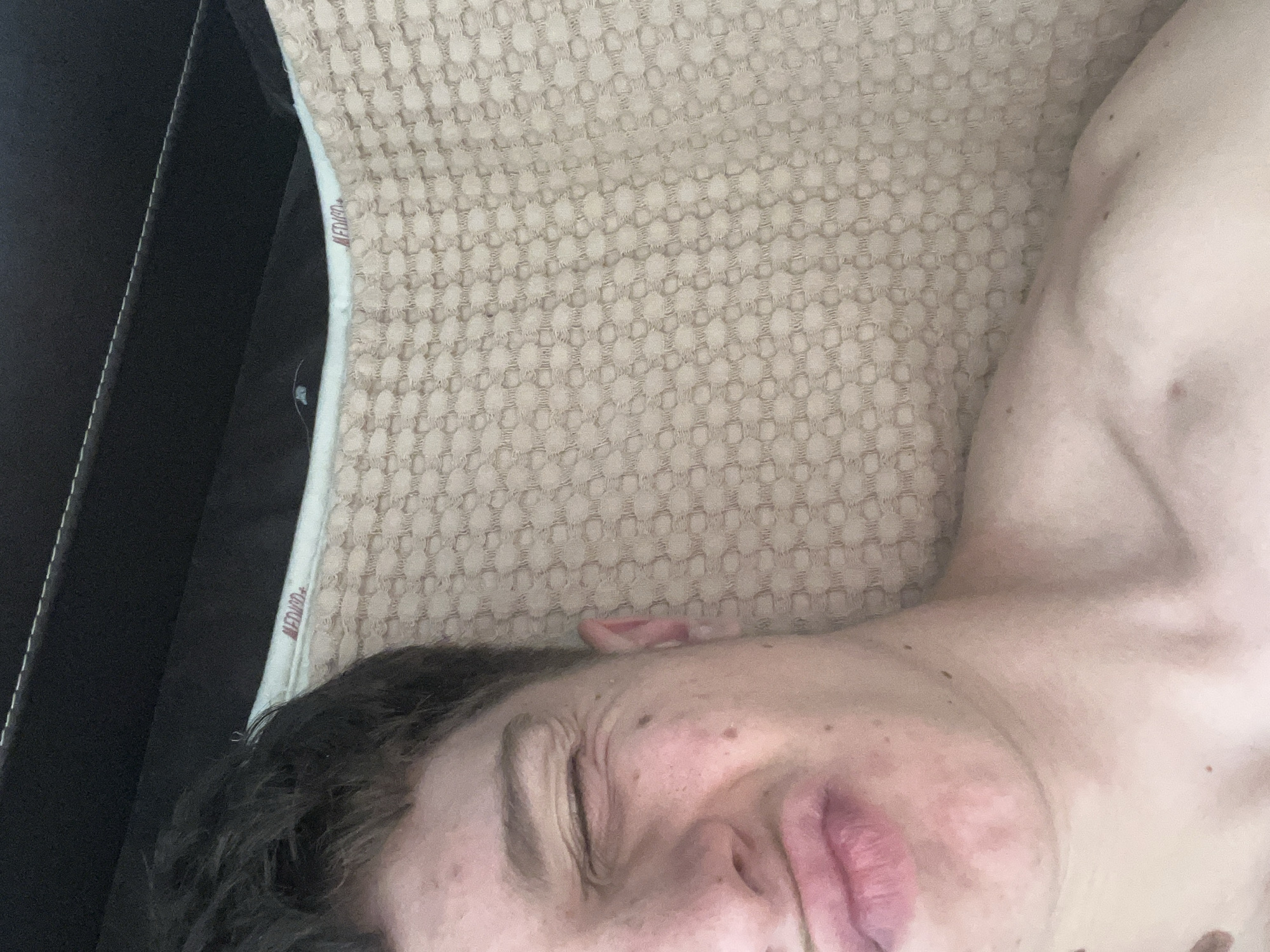 An overhead portrait captures a young individual, likely male, in the city of Zlatograd, Bulgaria, from the upper chest to the forehead, positioned vertically against a light-colored, textured fabric with a raised dot pattern. The individual's left eye is tightly closed, creating wrinkles at the temple, while their lips are pursed in a slight pout. Dark hair is visible on the top left. The person appears bare-chested, exposing the neck, collarbone, and upper shoulder. Behind the head, a light blue-grey fabric border is visible, featuring a red label with white text, partially decipherable as "MEDICD+" or "MEDICA+". Above this, a dark, possibly black, upholstered surface with visible stitching is present, suggesting a headboard or furniture. The scene is illuminated by soft, even light.