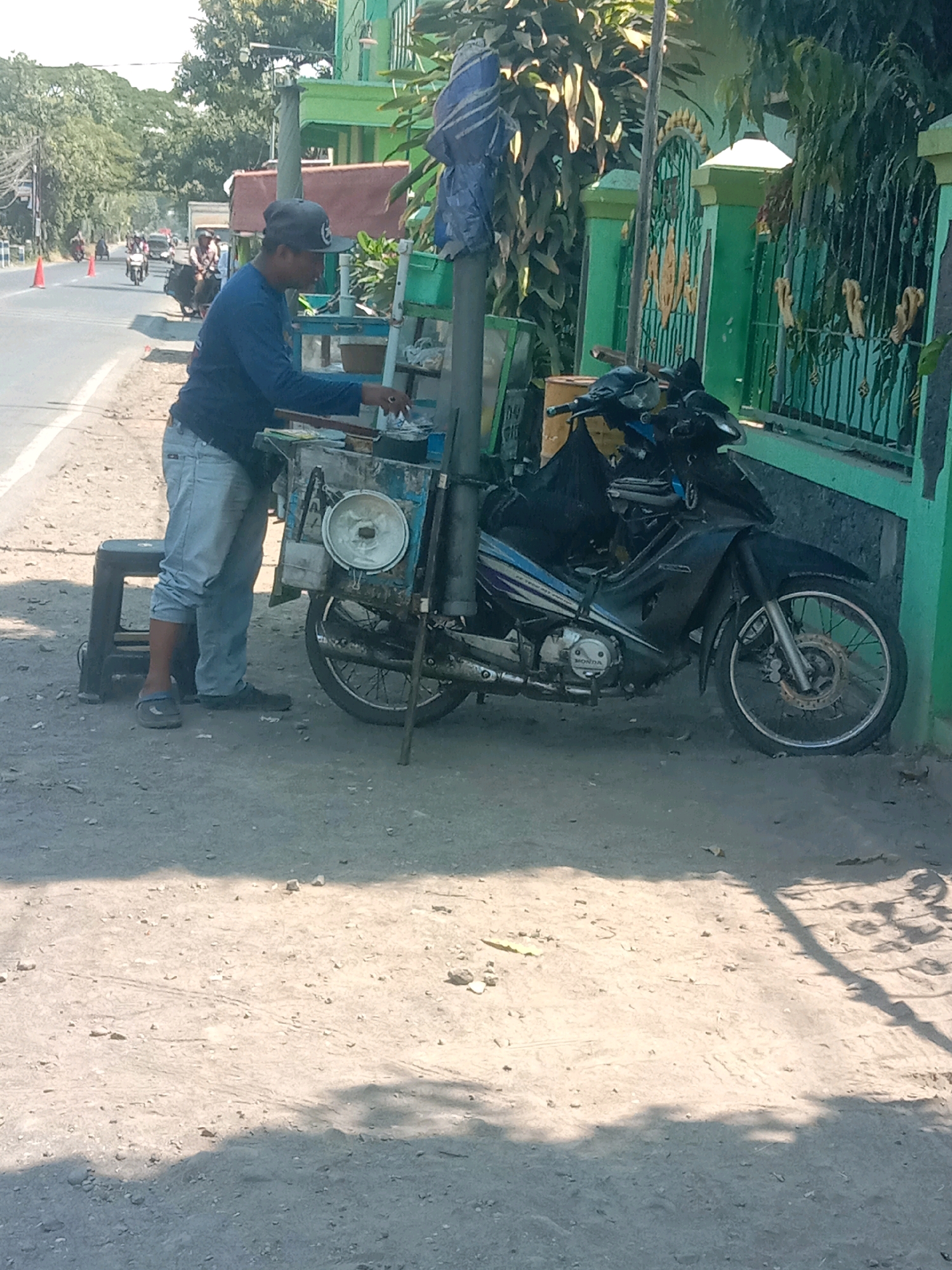A person wearing a blue long-sleeved shirt and light blue jeans stands next to a motorcycle that is parked on a dirt and asphalt road. The motorcycle is black and has a sidecar. The person is holding a small white object in their right hand and appears to be reaching into a box attached to the motorcycle. In the background, there is a green fence and a building. The scene is set in Karang Anyar, Indonesia, with traffic and trees visible in the distance. The lighting suggests it is daytime.