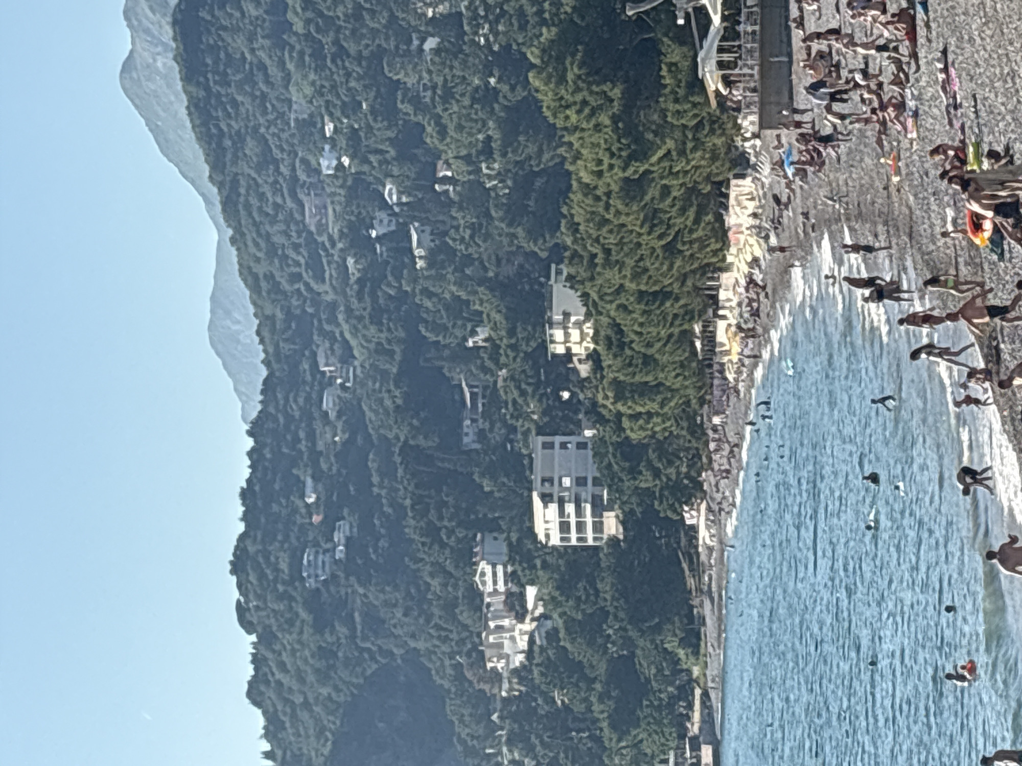 This image depicts a vibrant, sunny beach scene in Bar, Montenegro. A clear blue sky dominates the upper portion, with forested mountains rising in the background. Scattered among the dense green foliage on the mountainsides are numerous white and light-colored buildings, ranging from multi-story structures to individual houses.
In the midground, a line of lush green trees, possibly pines, separates the mountainous terrain from the beach. The foreground is occupied by a blue-turquoise sea with gentle waves lapping against the shore. Numerous individuals are visible in the water, swimming, wading, and standing, with one person on a red inflatable.
The beach itself is a busy, pebbled or rocky expanse, densely populated with people. Many are sunbathing on towels or sun loungers, some under small beach umbrellas. Others are walking, standing, or playing near the water's edge. Children are present among the adults. Along the rear edge of the beach, several white-roofed structures, possibly beach bars or restaurants with outdoor seating, are visible, furnished with white umbrellas and tables. A concrete or stone platform extends into the water on the far left. The scene is brightly lit by natural sunlight.