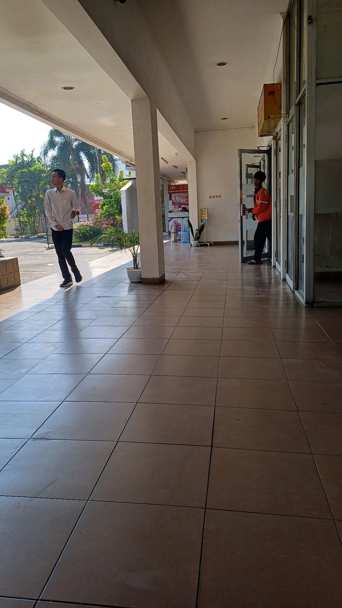 The image depicts an outdoor covered walkway in Pabelan, Indonesia, likely on a bright, sunny day given the strong shadows. The walkway is paved with large, square, tan tiles. On the left side of the walkway, a young man in a white long-sleeved shirt and dark pants is walking. He is casting a distinct shadow on the tiled floor. Further down the walkway, towards the right, another person is standing near an open glass door. This person is wearing an orange long-sleeved jacket and dark pants. To the right of the door, a building with glass-paneled walls is visible. Various potted plants are placed along the walkway. In the background, trees and parts of buildings can be seen. A yellow box, possibly a mailbox or signage, is mounted on the wall above the orange-jacketed person. There are also bottles of water visible inside the building through the glass doors. The architecture suggests a public building or commercial establishment.