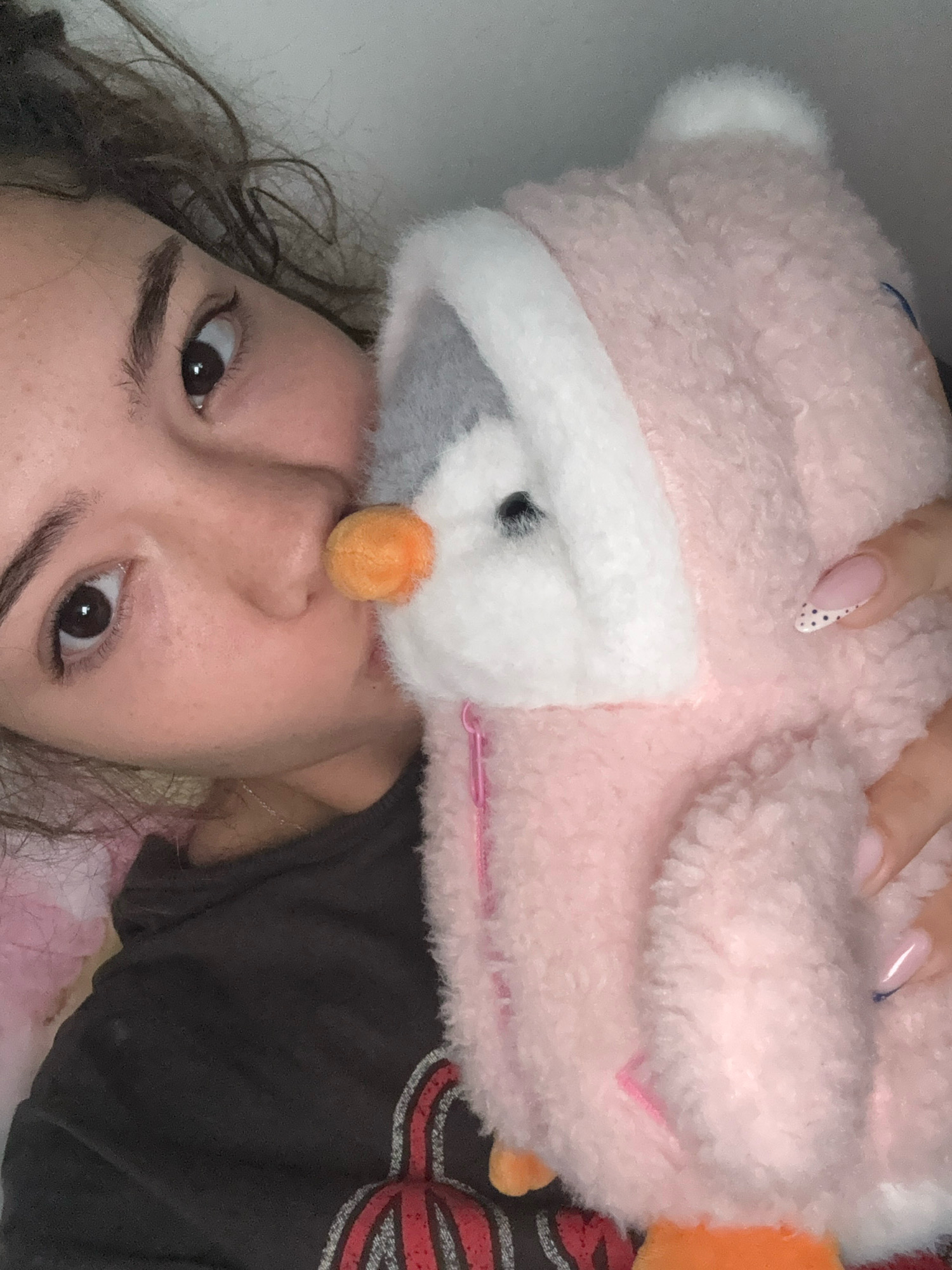 A close-up, angled, selfie-style shot shows a young woman with dark, curly hair, looking towards the camera with her right eye more prominent. She is holding a plush toy, possibly a penguin, which is predominantly pink with white and gray accents. The woman's left cheek is pressed against the toy. She is wearing a dark gray t-shirt with a red and black graphic design. Her fingernails are visible and appear to have a French manicure with black polka dots. The lighting is soft and diffused, suggesting an indoor environment. The location is Unknown, Unknown.