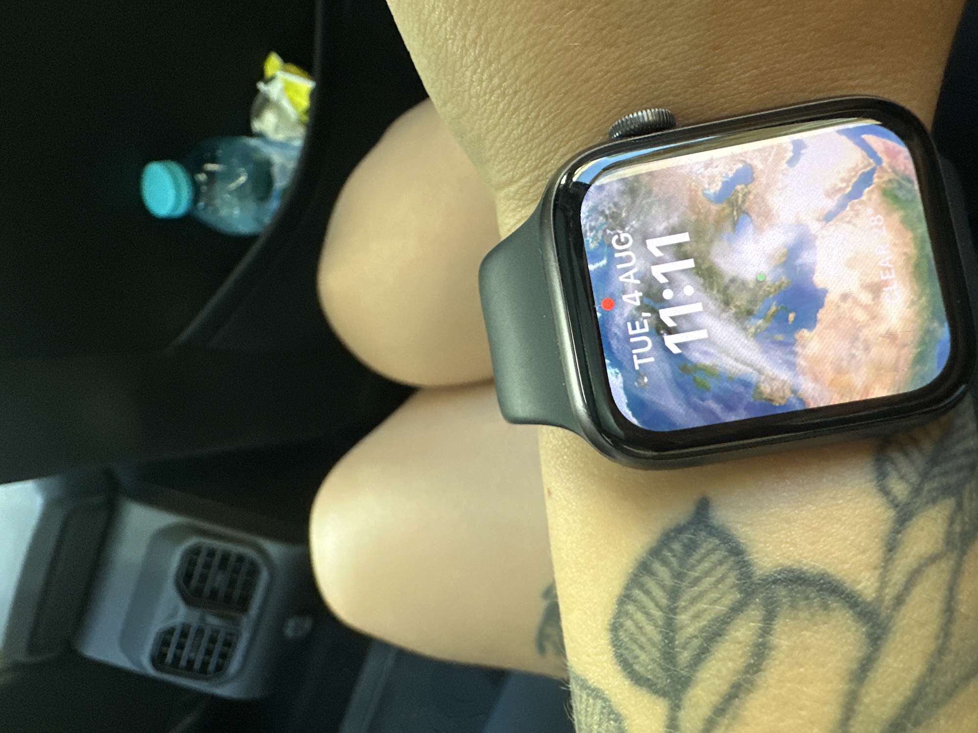 The image shows a close-up view of a person's wrist wearing an Apple Watch. The watch face displays the date "TUE, 4 AUG" and the time "11:11" with a world map background. The person's skin has a tattoo on their arm, and their legs are visible in the background, along with car interior elements like air vents and a water bottle. There is no information in the image that can identify the location as Plovdiv, Bulgaria.