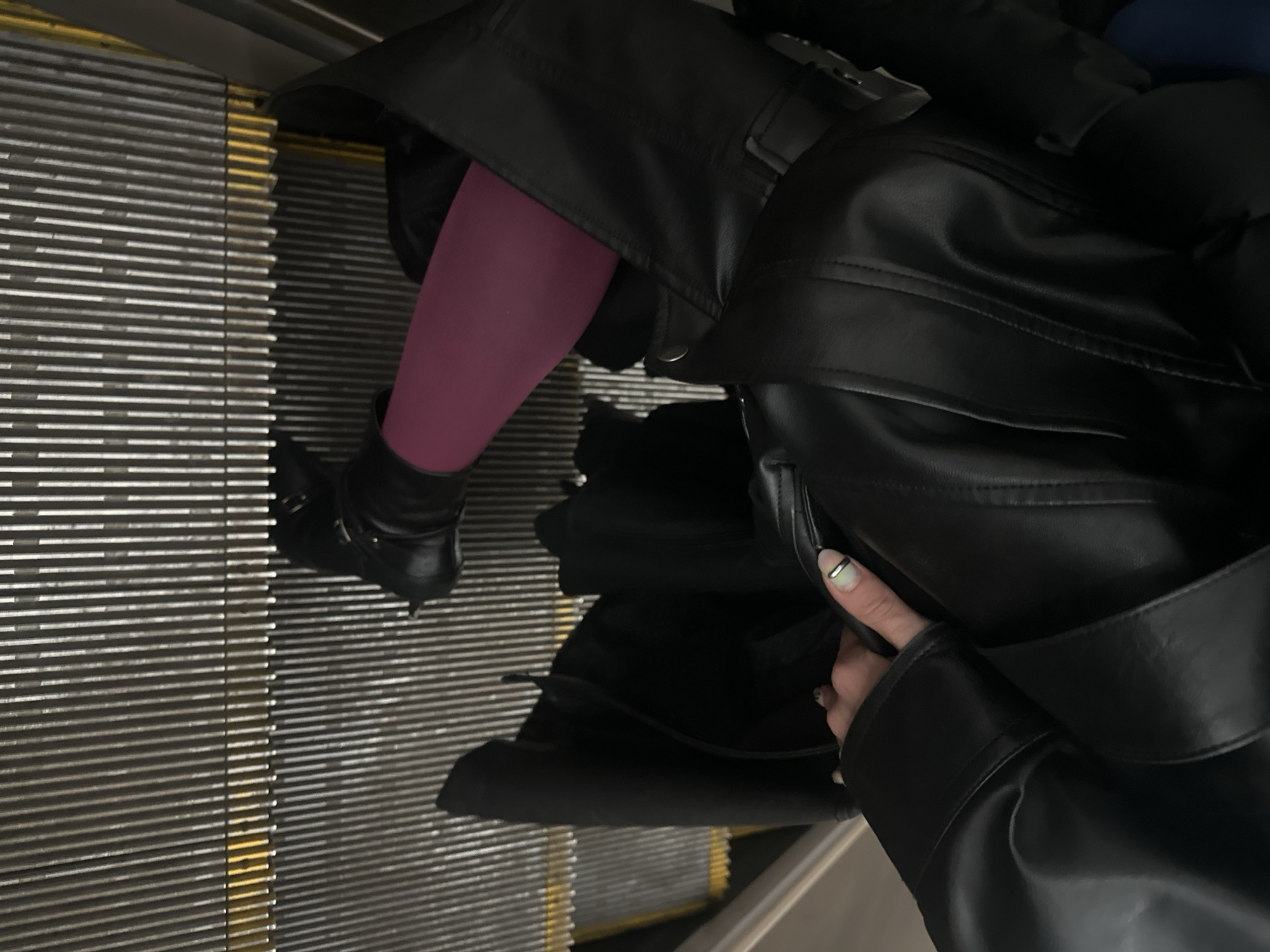The image shows a person's legs and torso from a first-person perspective, as if the photographer is looking down. The person is wearing a black leather jacket, magenta tights, and black boots. They are standing on an escalator with a metallic grate-like surface. The escalator has yellow safety markings. The overall lighting is dim. The image provides no information about the location in Moscow, Russia.
