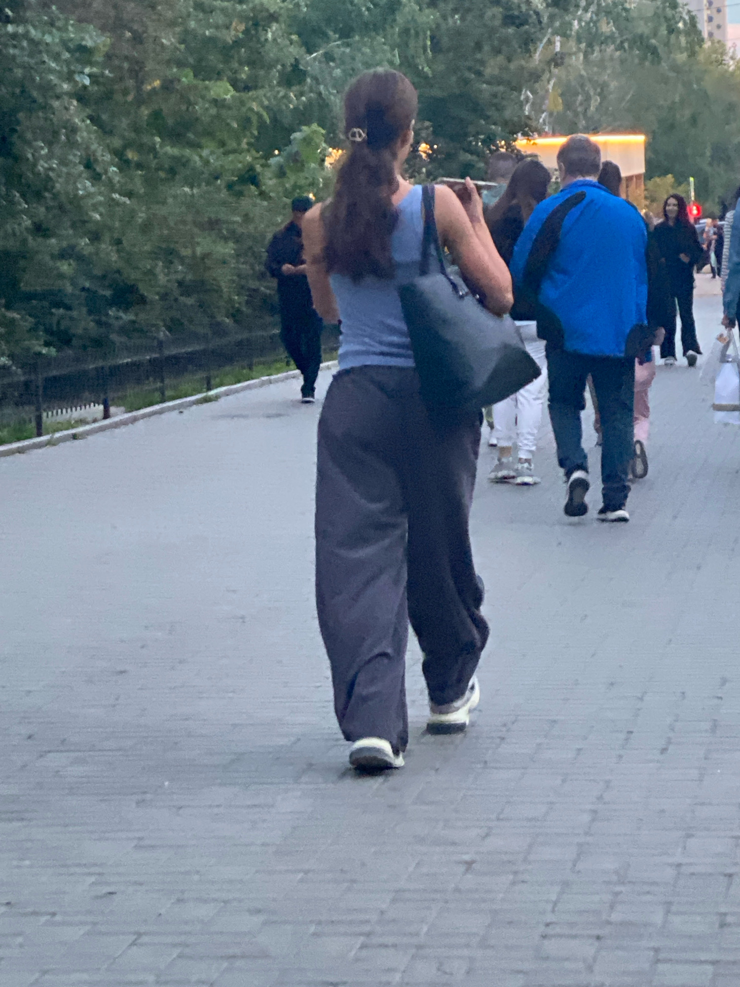 A person with dark hair is walking away from the camera on a paved walkway, wearing a light blue tank top and dark grey pants. They are carrying a dark tote bag over their left shoulder. Behind them, several other people are walking in the same direction. A person in a bright blue jacket is visible, as are others in darker clothing. The walkway is bordered by a black fence and lush green trees. In the background, buildings with illuminated signs are present, suggesting an urban environment. The paving stones of the walkway are a neutral gray.