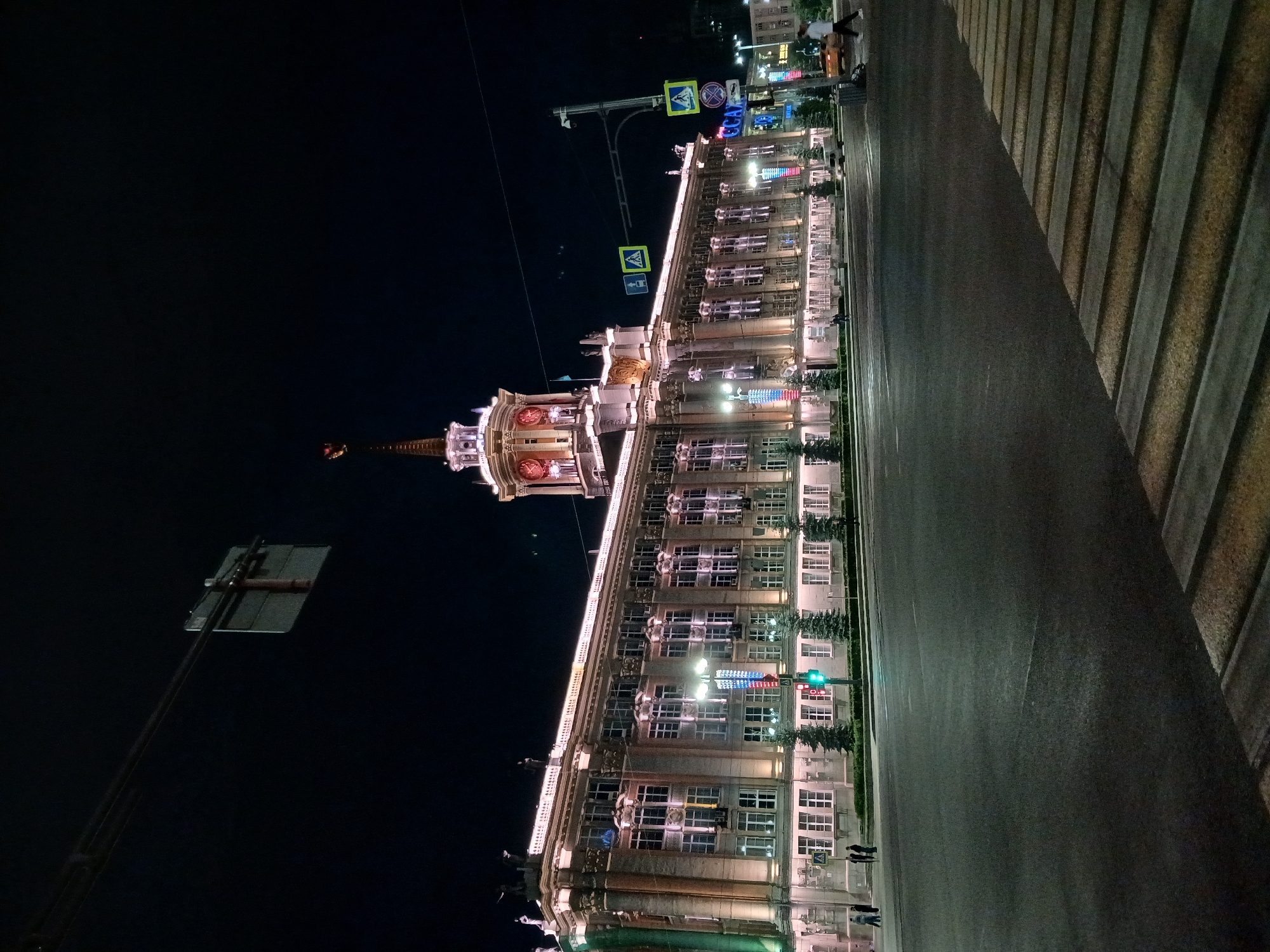 This is a night time, wide angle, ground-level outdoor photograph of the Yekaterinburg City Administration Building, located in Yekaterinburg, Russia. The building is illuminated with bright white and pink lights, highlighting its architectural features. The building is large, with multiple floors and many windows. A prominent clock tower with red clock faces is visible on the right side of the building. A flag is flying from the top of the tower.
In the foreground, there is a wide, multi-lane asphalt road with a pedestrian crossing. The road surface is dark and reflective due to the lighting. Traffic lights are visible on the road, with a green light illuminated. Several street lights are positioned along the sidewalk in front of the building.
There are a few pedestrians walking on the sidewalk in front of the building and near the crossing. A yellow car is visible in the distance on the right side of the image. Several decorative trees are planted along the sidewalk. The sky is dark, indicating nighttime. Overhead, a traffic signal hangs from wires.