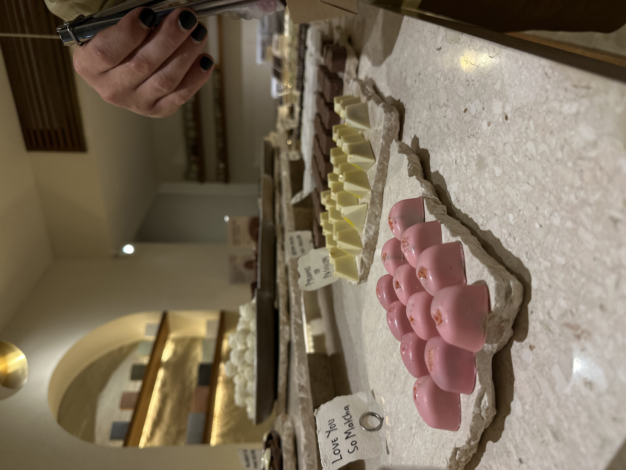 The image shows a display of chocolates in a store, likely a chocolatier. A hand holding metal tongs is visible in the upper left, presumably to select chocolates. The chocolates are arranged on rough, light-colored stone slabs, with small handwritten labels identifying the flavors. The labels visible read "Love You So Matcha" and "Pyramid of Passion." The chocolates include heart-shaped pink ones with a speckled appearance, white pyramid-shaped chocolates, and some brown square chocolates further down the display.
In the background, a curved wall feature with recessed shelves is visible, adding to the ambiance of the store. The specific location within the city of Unknown, Unknown, is not discernible from the image.