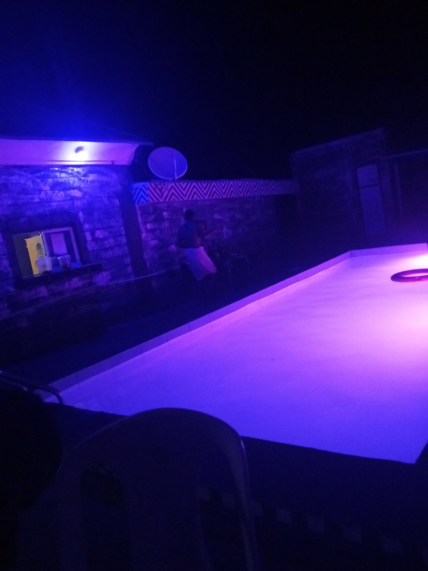 The scene shows an outdoor pool area at night, likely in Lokoja, Nigeria. The pool is illuminated with purple light. A person wearing a white top stands in the background. There's a building with a satellite dish and a window in the background. There's an object in the foreground that resembles a chair.