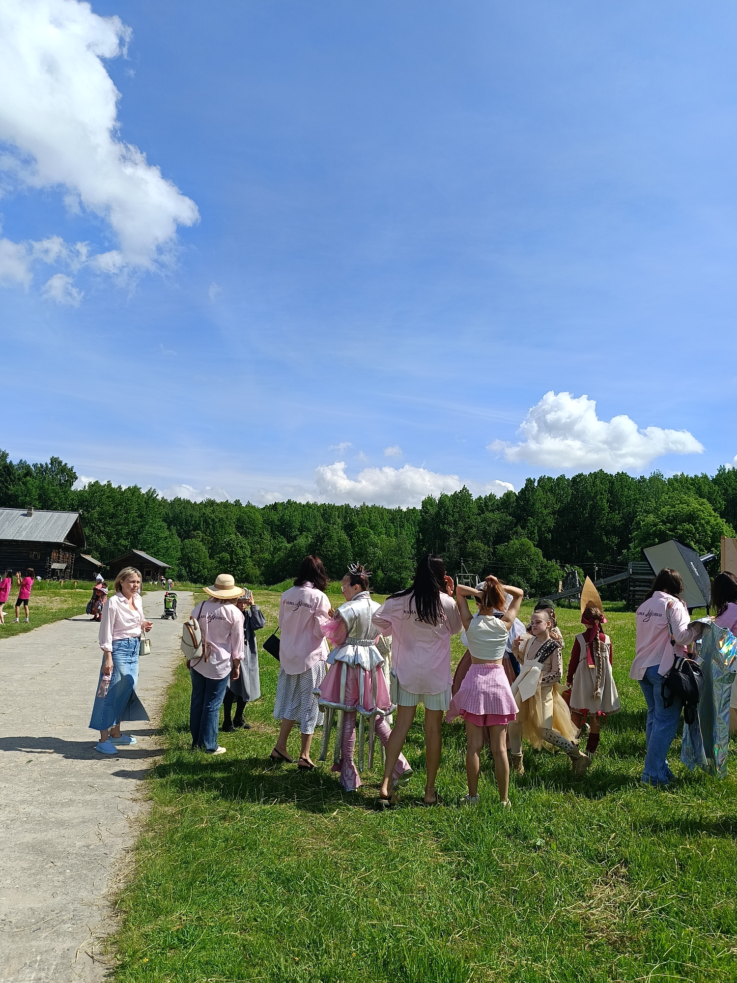 In an outdoor setting with a blue sky dotted with white clouds, a group of people, mostly women and girls, are gathered on a grassy area next to a paved path. Several individuals are wearing matching pink shirts with text that appears to read "МАМА ЛЮБОВЬ" (Mama Love). Some are wearing costumes or brightly colored outfits. In the background, there are wooden buildings and a dense forest. The scene appears to be at an event or gathering. The location is identified as Popovka, Russia.