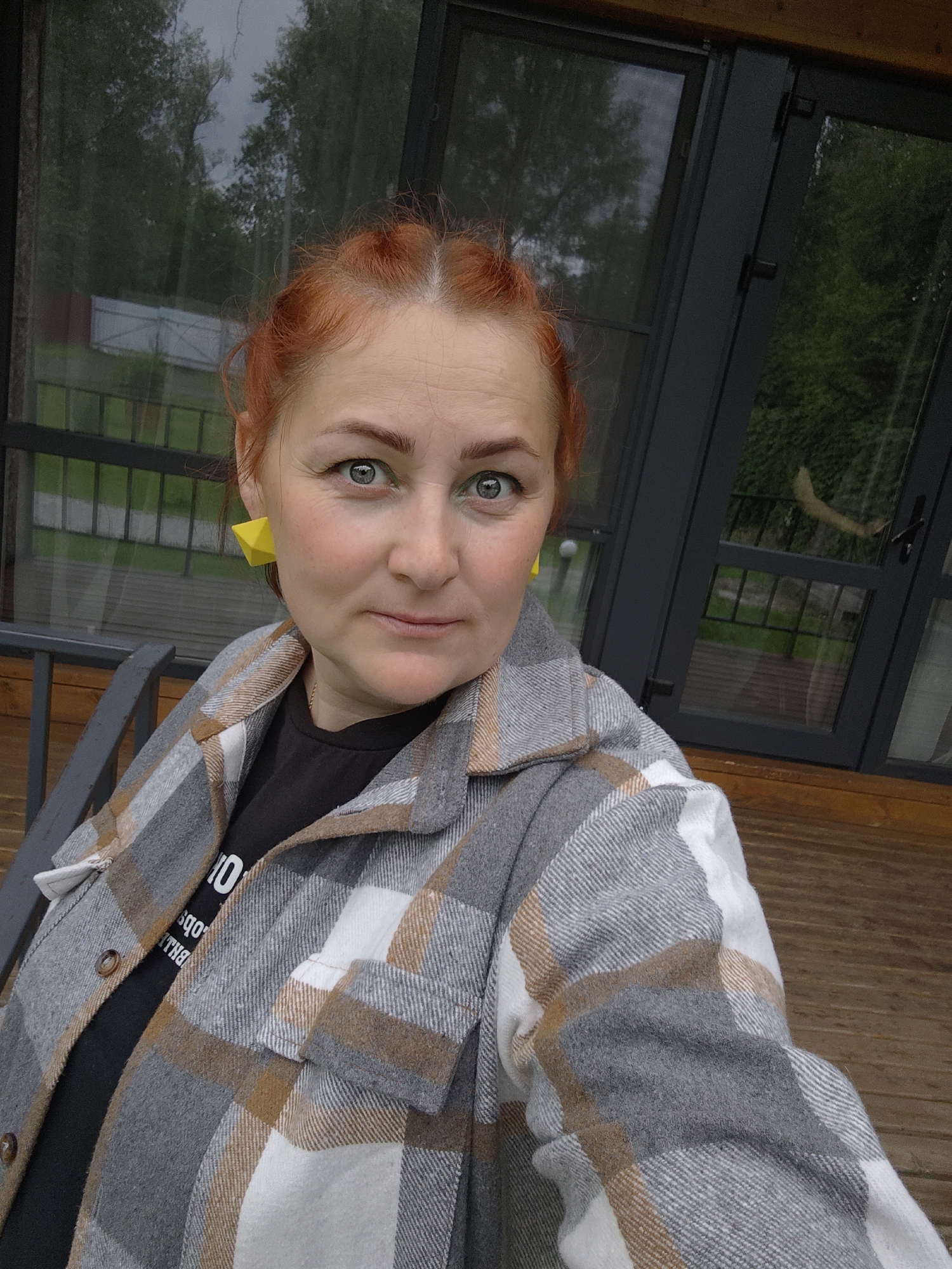 An adult female is depicted in a medium-close-up selfie, positioned slightly off-center to the left, looking directly at the camera. She has red hair, partially styled in visible braids or buns at the top of her head, and fair skin. Her eyes are light grey-green, with a visible green eyeliner or eyeshadow on the lower lid. Her facial expression is neutral to slightly smiling. She is wearing large, geometric, bright yellow stud earrings. Her attire consists of a grey, brown, and white plaid flannel shirt worn open over a black t-shirt. The t-shirt has white text, partially visible as "10 qod obc THL" (likely inverted/reflected text).
The background indicates an outdoor setting, likely a wooden porch or balcony, with a brown wooden floor and a dark grey metal railing in the lower left. Behind her are large windows with dark grey frames, reflecting overcast skies and green trees. The reflections suggest a natural, possibly wooded, environment. The overall scene takes place in Malyi Bukor, Russia.