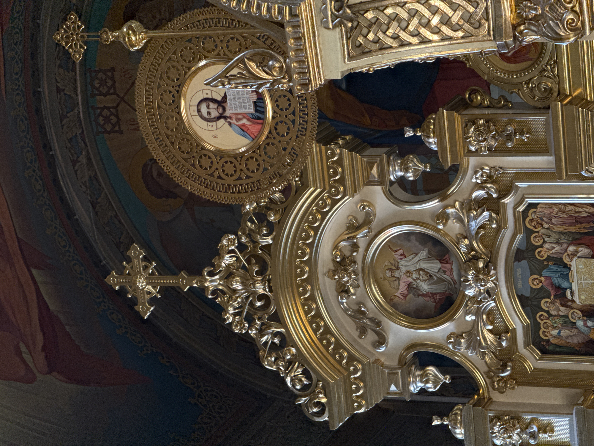 This image shows the interior of an Orthodox church, likely the iconostasis, a wall of icons and religious imagery. The scene is dominated by ornate golden architectural elements and religious paintings. In the foreground, a large, intricately carved golden structure, possibly part of the iconostasis, is visible. It features decorative motifs, including floral patterns and scrollwork.
Above the golden structure, several circular icons are displayed. One prominent icon, framed by a decorative golden circle, depicts Jesus Christ holding a book and making a gesture of blessing. To its left, another circular icon shows God the Father holding the infant Jesus, with both figures in white and pink robes.
Below the central golden structure, another religious painting is visible. This painting appears to depict a scene from the Last Supper, with Jesus and his disciples gathered around a table. The figures are rendered in traditional Orthodox style, with halos around their heads.
The background of the image shows painted murals on the ceiling or upper walls of the church, with large figures in red and blue robes. The overall impression is one of opulence and religious devotion, characterized by rich gold embellishments and devotional imagery typical of Orthodox Christian art. The lighting is somewhat dim, with highlights reflecting off the polished gold surfaces.