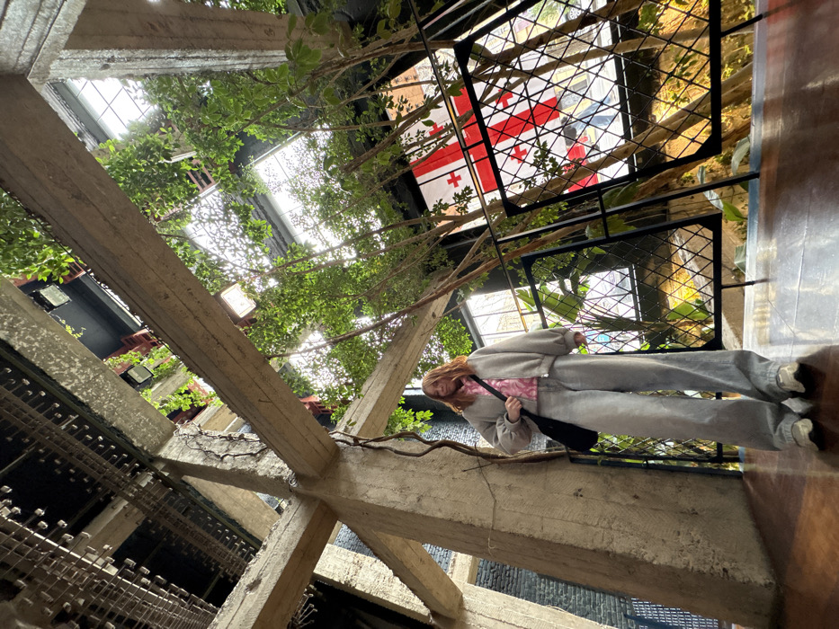 The image displays the interior of a multi-story building in Tbilisi, Georgia, specifically identified as an atrium resembling that of Rooms Hotel Tbilisi. The environment features a raw brutalist aesthetic with prominent exposed concrete beams forming a structural grid that extends vertically and overhead. Cracks and exposed rebar are visible on some concrete elements, with thin, dried vines or roots clinging to them. Numerous small, leafy trees and green plants are strategically integrated throughout the space, growing upwards and from various levels, creating a dense natural canopy against the concrete and overhead structures.
A female individual stands in the lower-left foreground on a polished dark wood floor. She has shoulder-length reddish-brown hair and is dressed in a light grey oversized bomber jacket, a pink patterned top, wide-leg light grey jeans, and light-colored athletic shoes. She carries a black shoulder bag on her left shoulder. Her posture is relaxed, facing slightly to the right of the frame.
Through a large window on the right side of the image, the red and white flag of Georgia is prominently visible, indicating the country. Outside the window, a blurry street or building exterior is partially discernible. A black metal railing with a diamond-grid pattern runs along the right side of the viewing platform. Natural light streams into the space from the large windows, brightly illuminating the foliage and upper sections of the building.