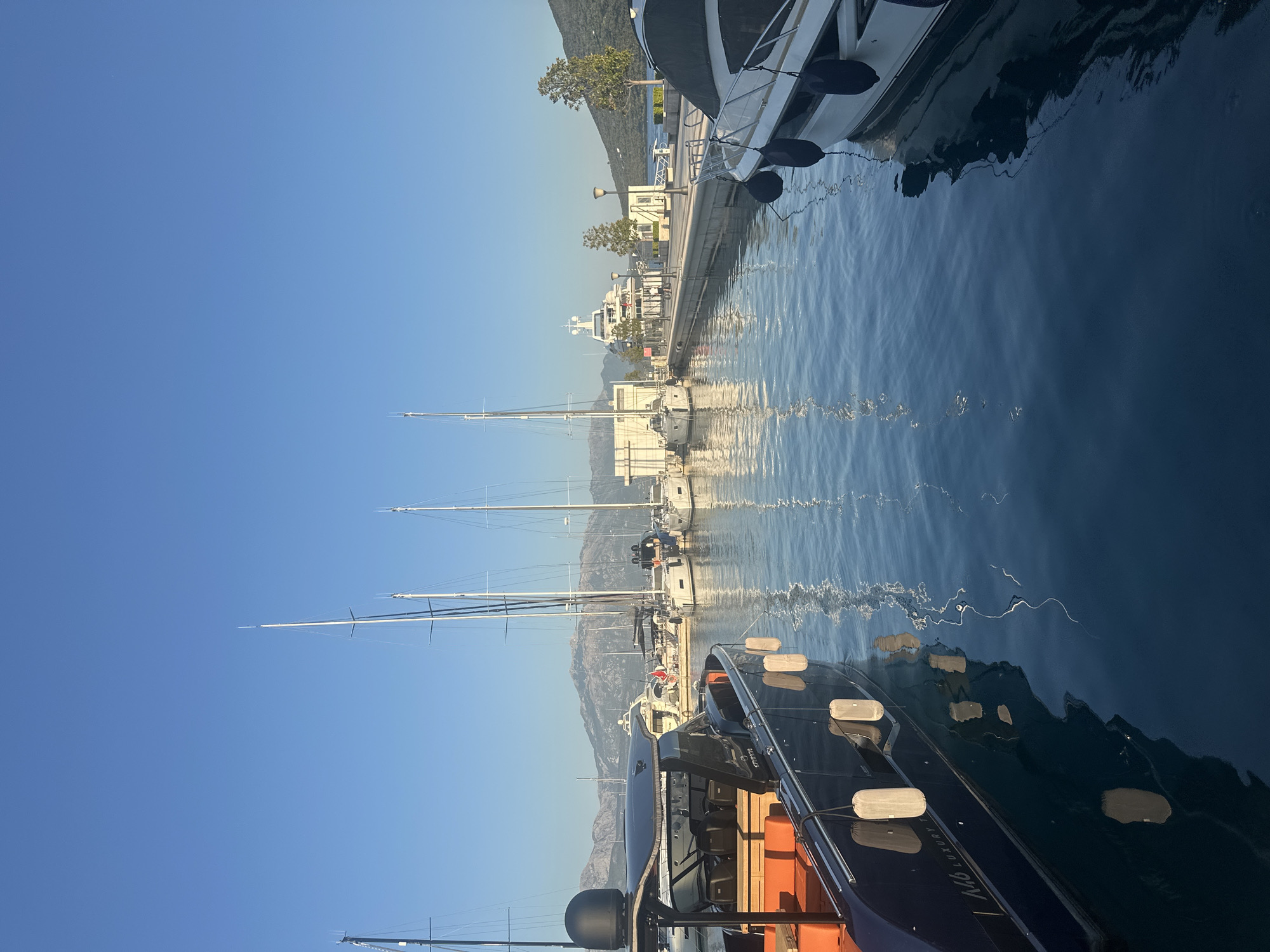 The image depicts a marina in Herceg Novi, Montenegro, under a clear blue sky. In the foreground, two boats are docked. The boat on the left is a dark-hulled yacht with orange seating visible. It has several white fenders hanging along its side. Text on its hull reads "M46 LUXURY". The boat on the right is a white motor yacht with dark blue fenders. Several sailboats with tall masts are moored in the middle ground, their reflections visible in the calm, deep blue water. In the background, a large white superyacht is docked at a pier lined with lampposts and trees. Mountains form the backdrop to the scene. Two individuals are standing on the pier near the superyacht. The lighting suggests it is morning or late afternoon.